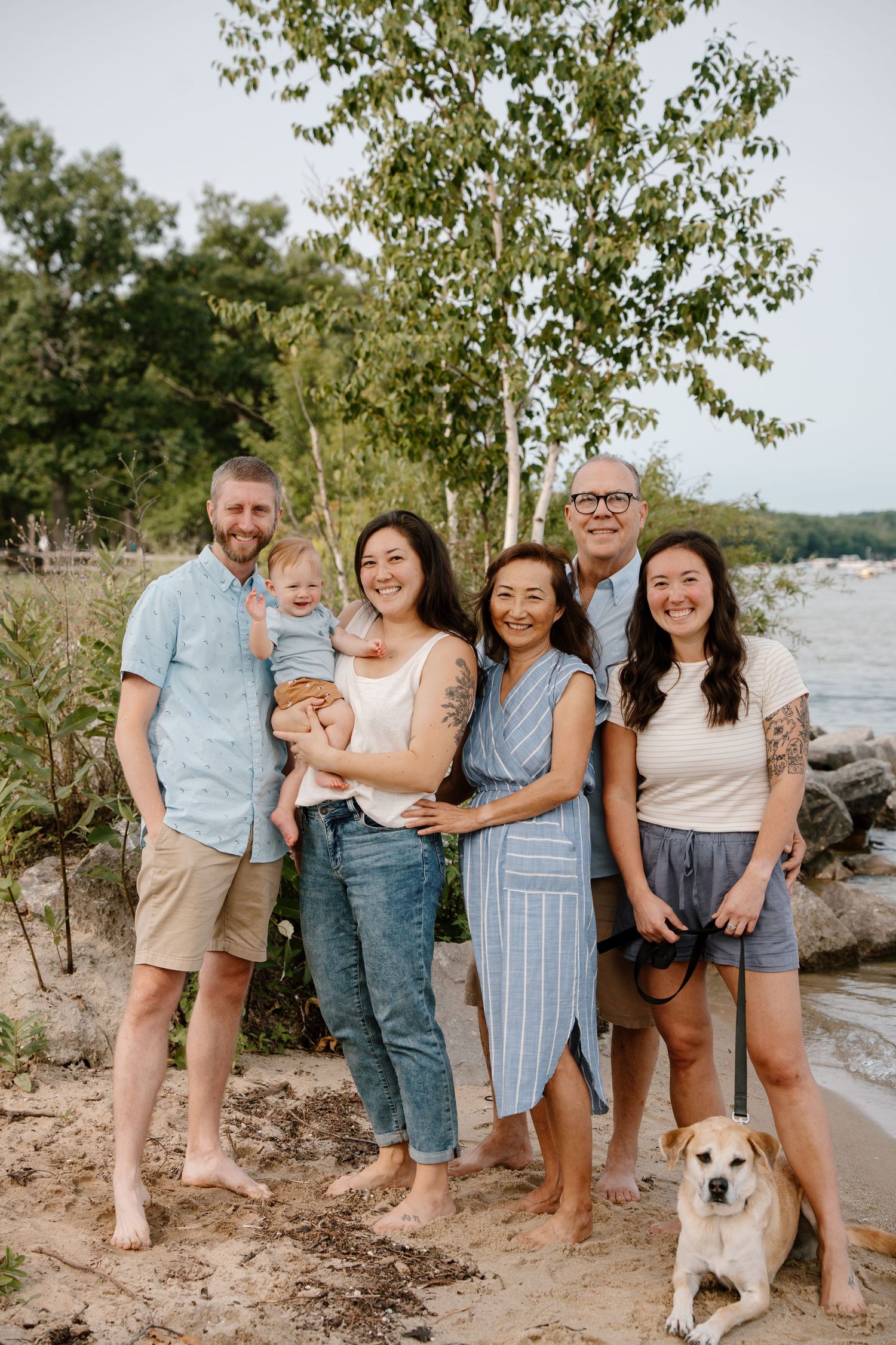 A joyful family of six poses on a sandy beach with a dog, trees, and water in the background.