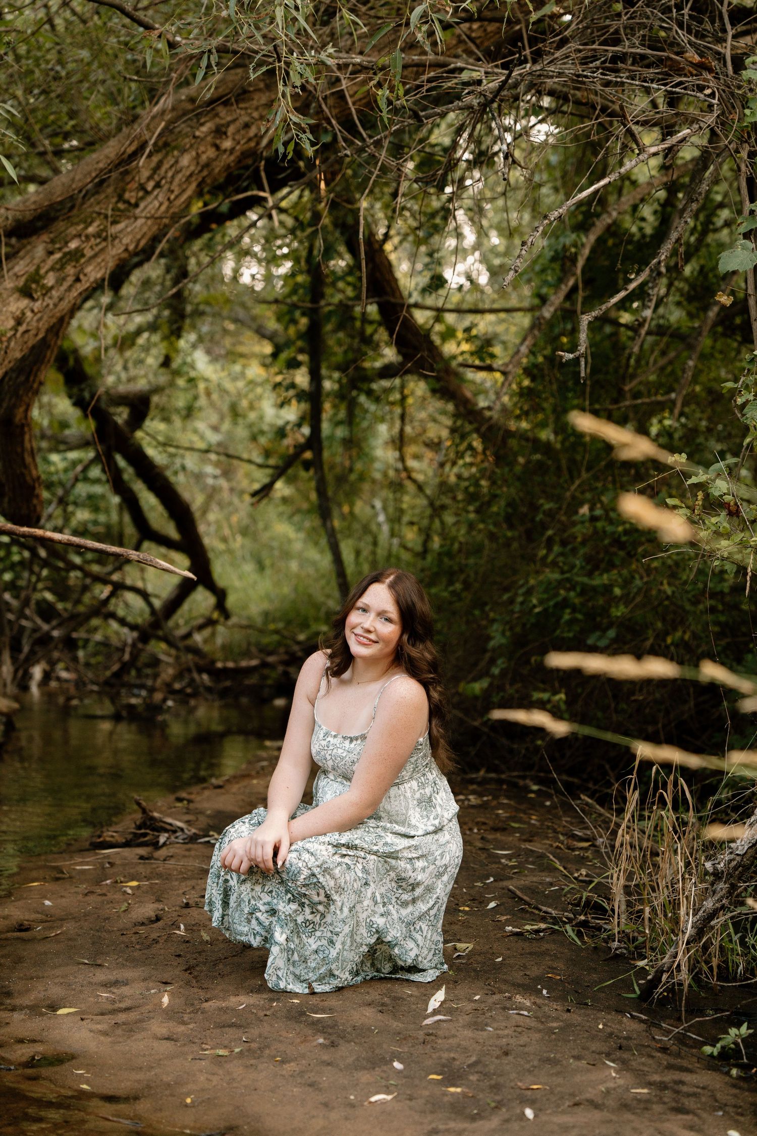 Young woman in floral maxi dress sitting by a creek in a lush forest setting, smiling naturally.