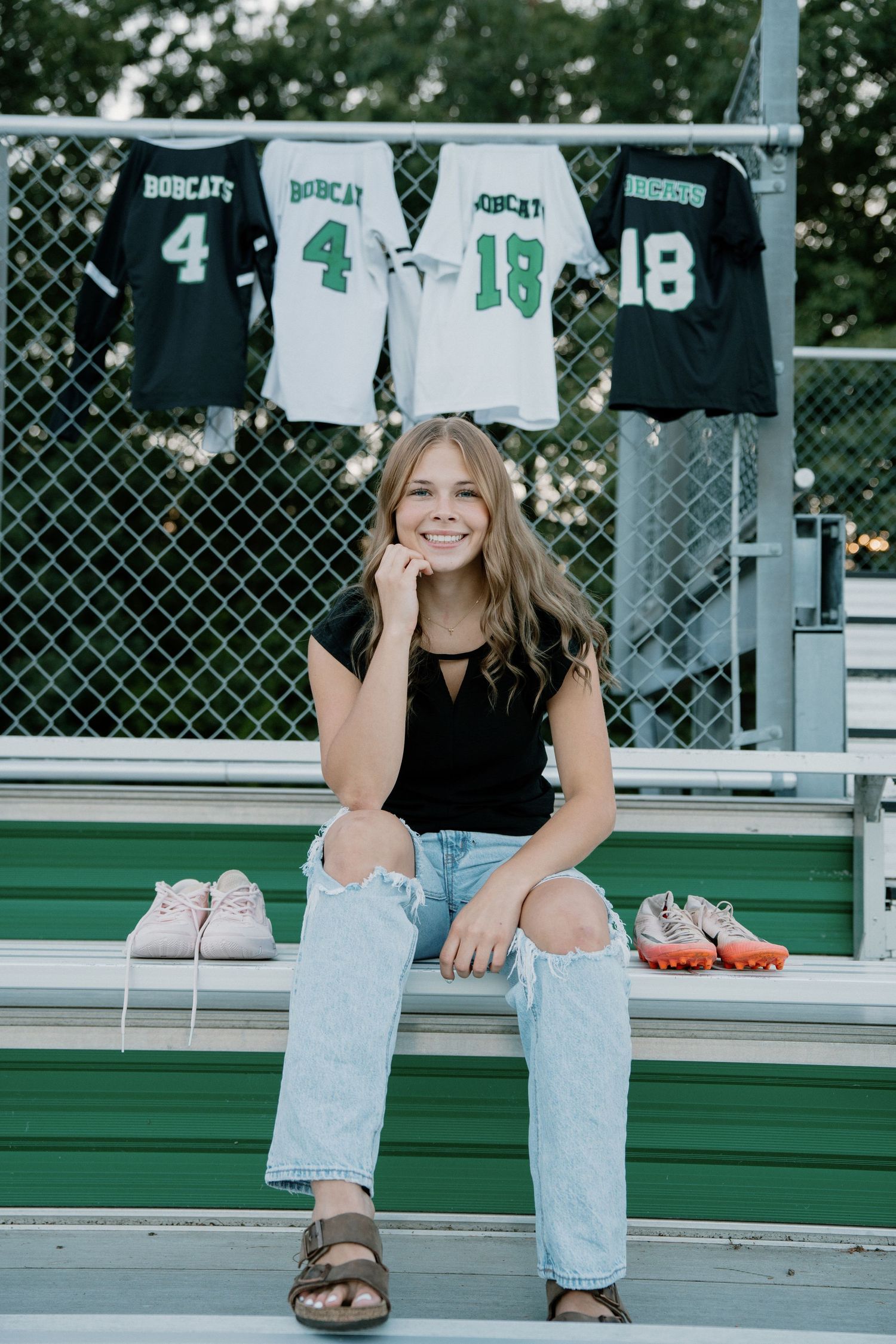 Smiling teen girl sits on bleachers beside Bobcats jerseys numbers 4 and 18 hanging on a chain-link fence.