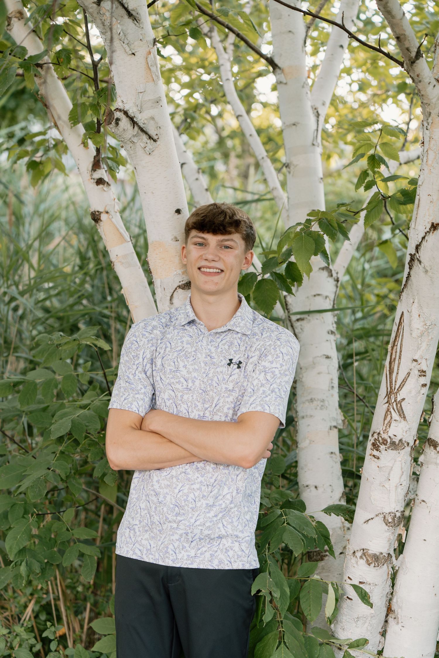 Smiling teenage boy in Under Armour polo stands with arms crossed among white birch trees outdoors.
