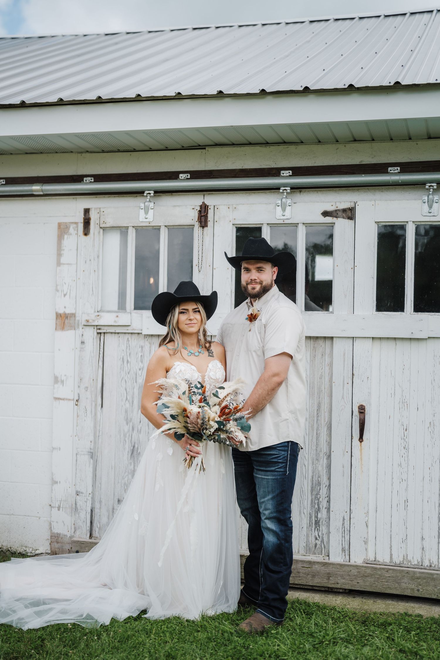 Western-themed wedding couple poses in front of rustic white barn.