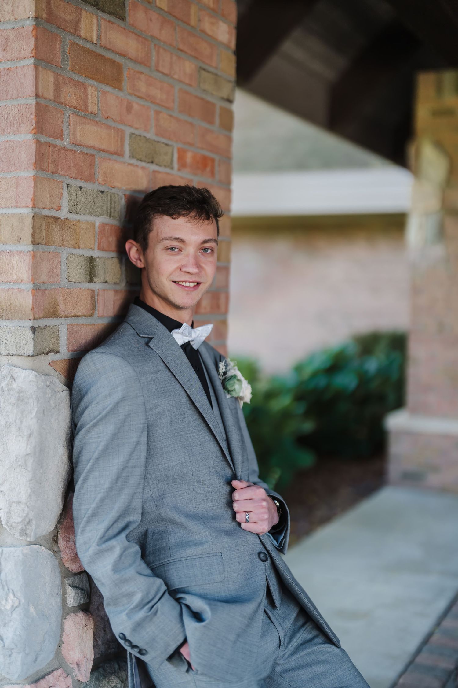 Person in grey suit and bowtie leans against brick wall with boutonniere.