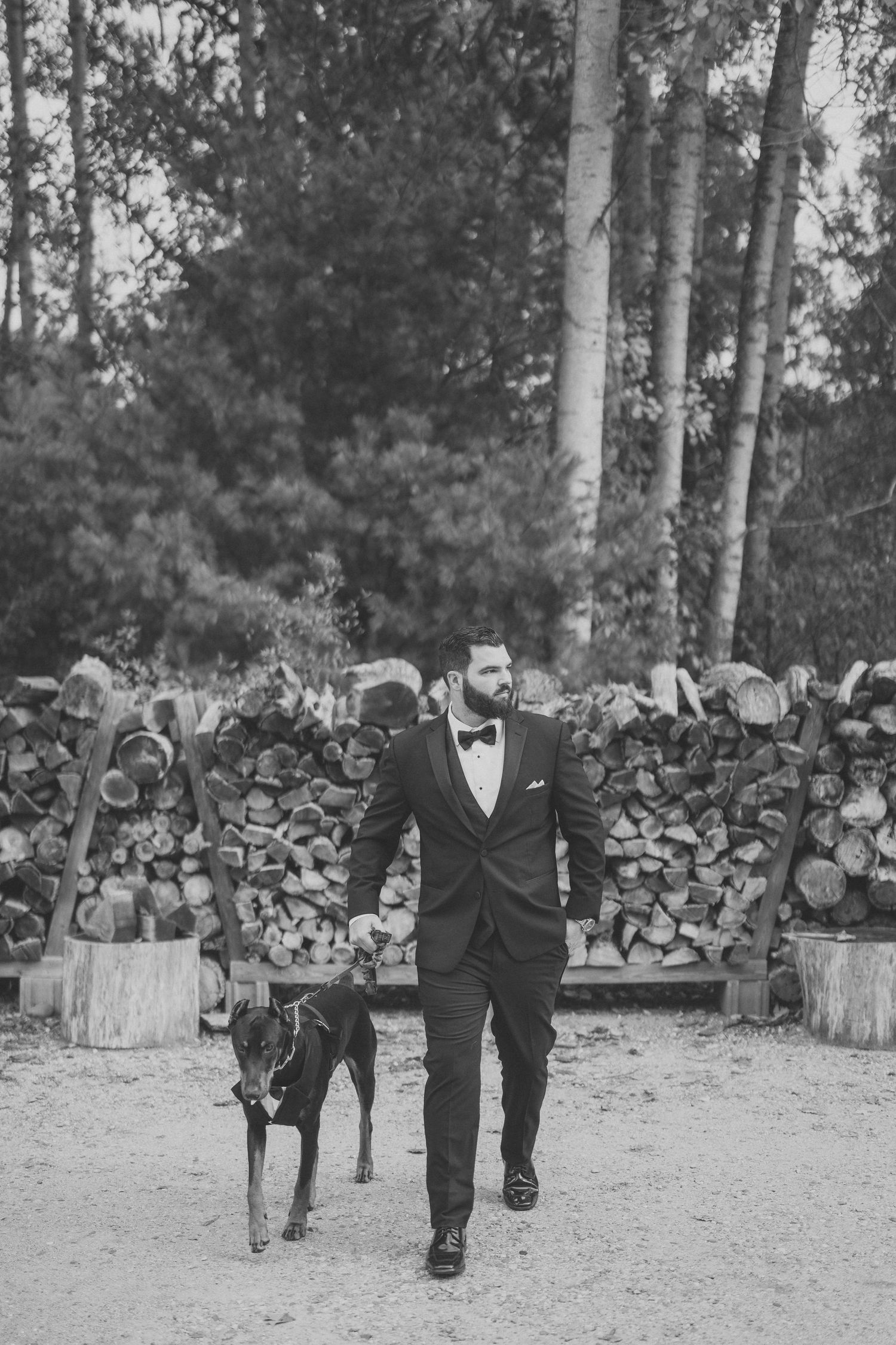 Black and white wedding photo of groomsmen walking along a rustic wood pile backdrop.