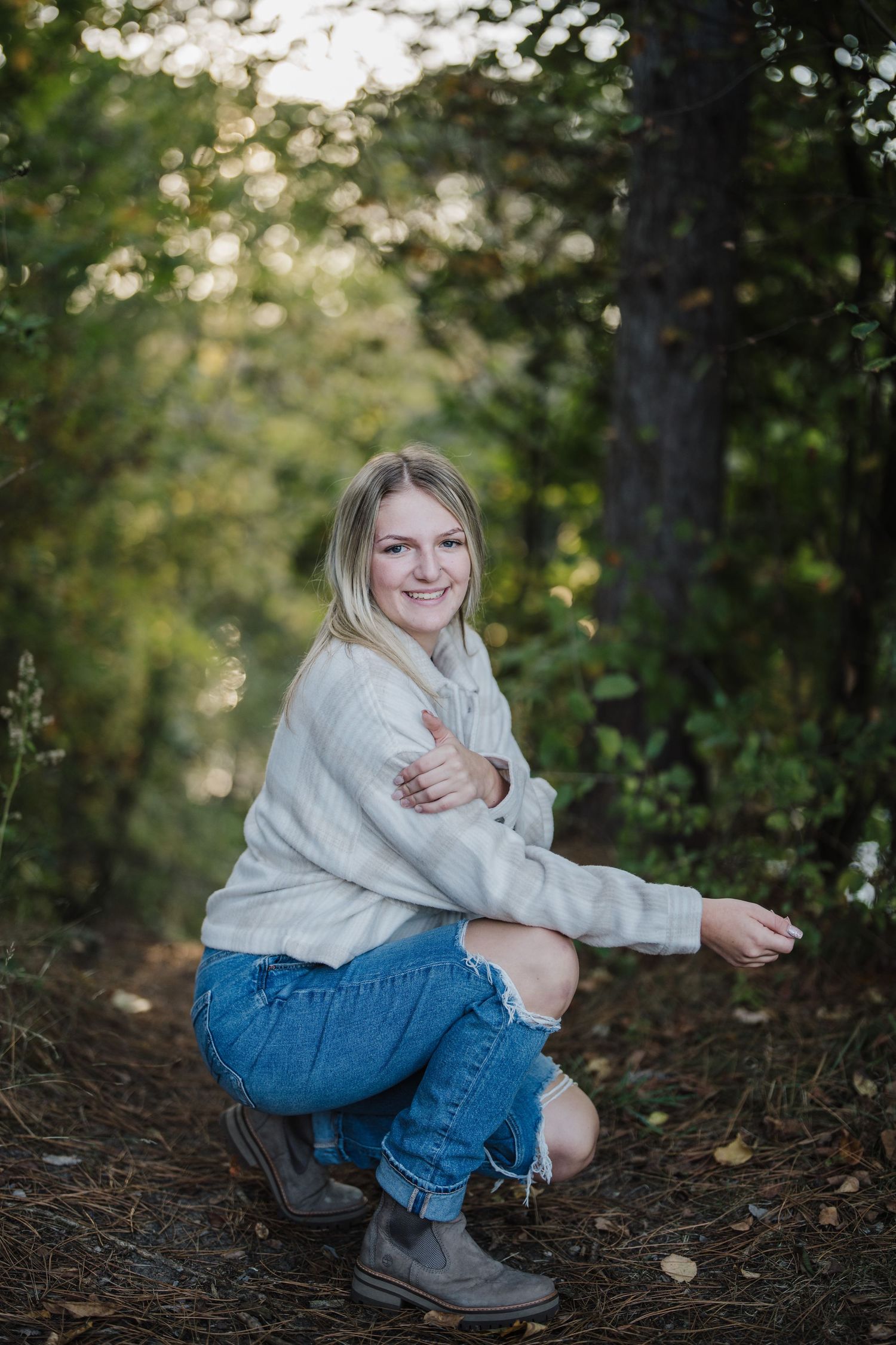 Casual outdoor photoshoot featuring someone in a cream sweater and denim sitting among fallen leaves and trees.