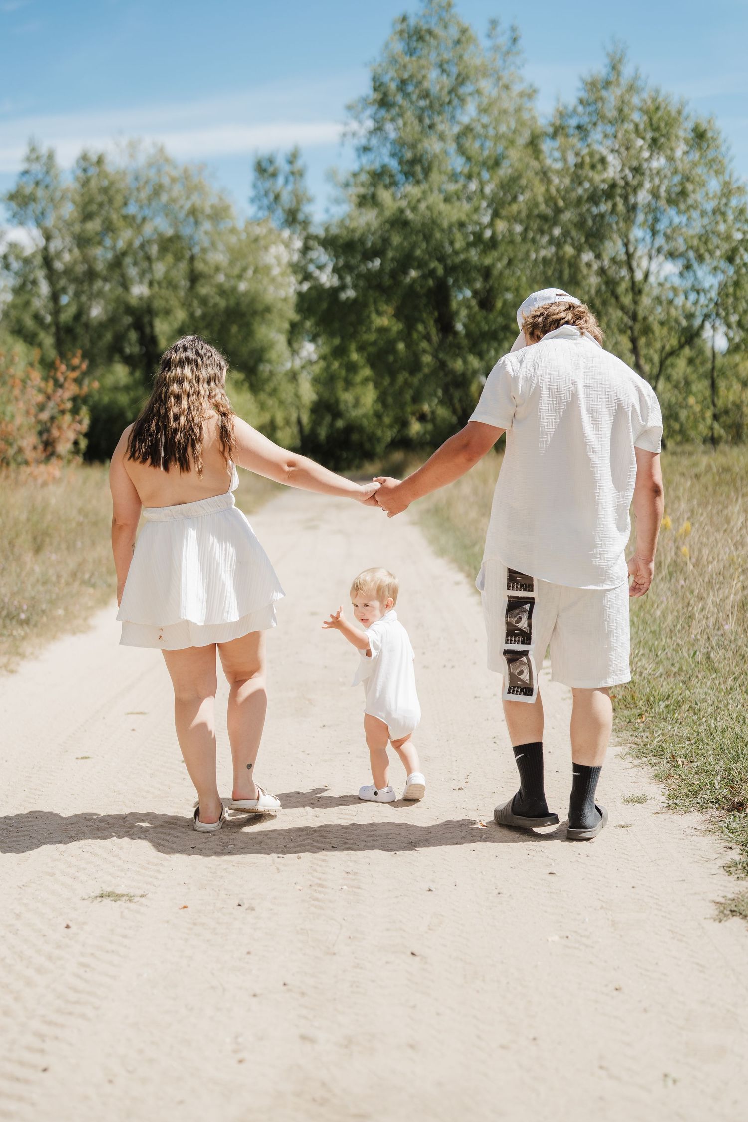 A family wearing white outfits walks together on a sandy path surrounded by green foliage on a sunny day.