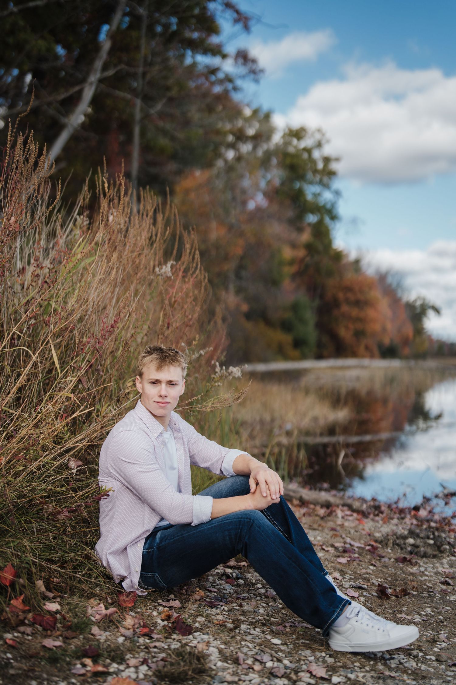 A casual outdoor photoshoot on a rocky shoreline with autumn foliage and water in the background.