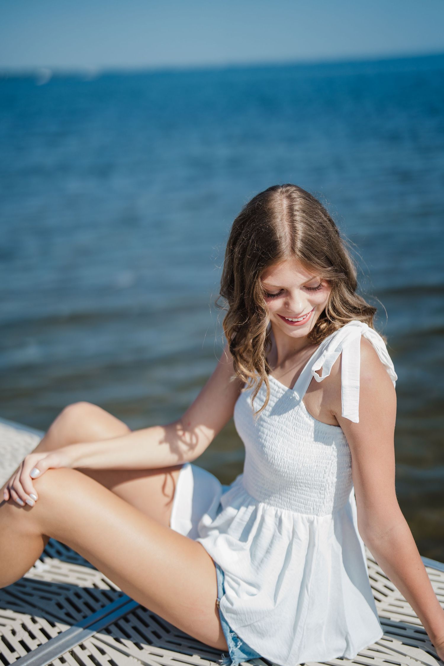 A summery beach portrait featuring someone in a white sundress sitting on a blanket by the ocean.