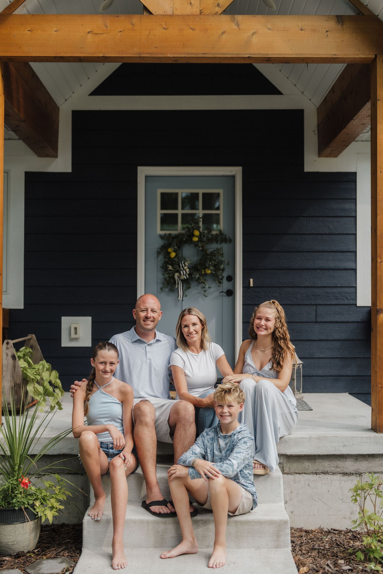 Happy family of five posing on the front porch steps of a navy blue home on a sunny day.