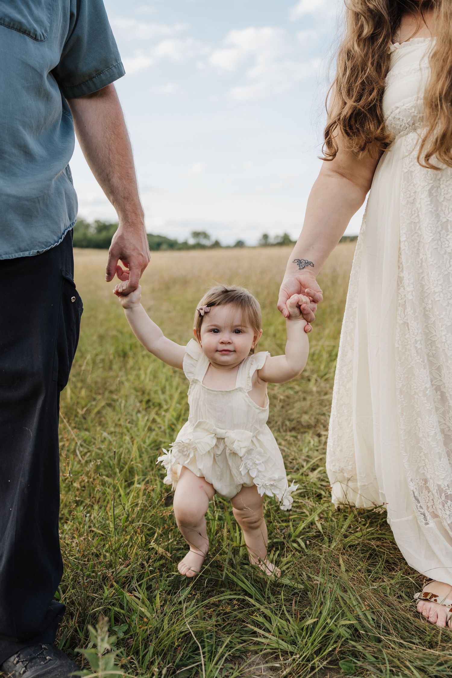 A baby in a cream romper holds parents hands while standing in a green field on a cloudy day.