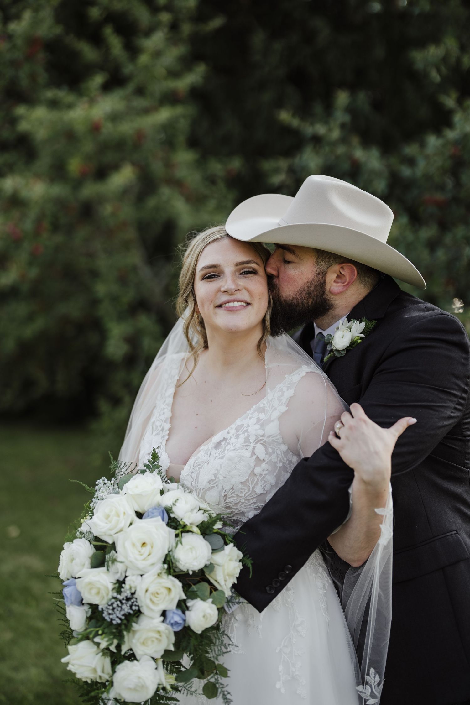 Wedding couple shares intimate moment outdoors with white floral bouquet and cowboy hat details.