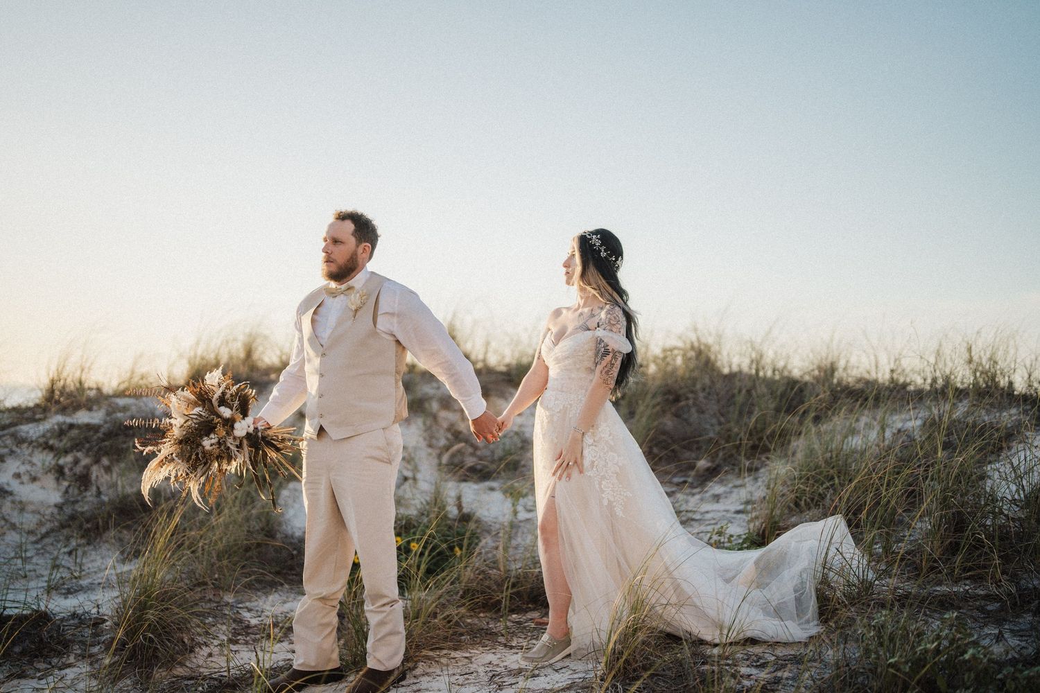 A couple in wedding attire holds hands on a sandy beach dune during sunset while the bride holds a dried floral bouquet.