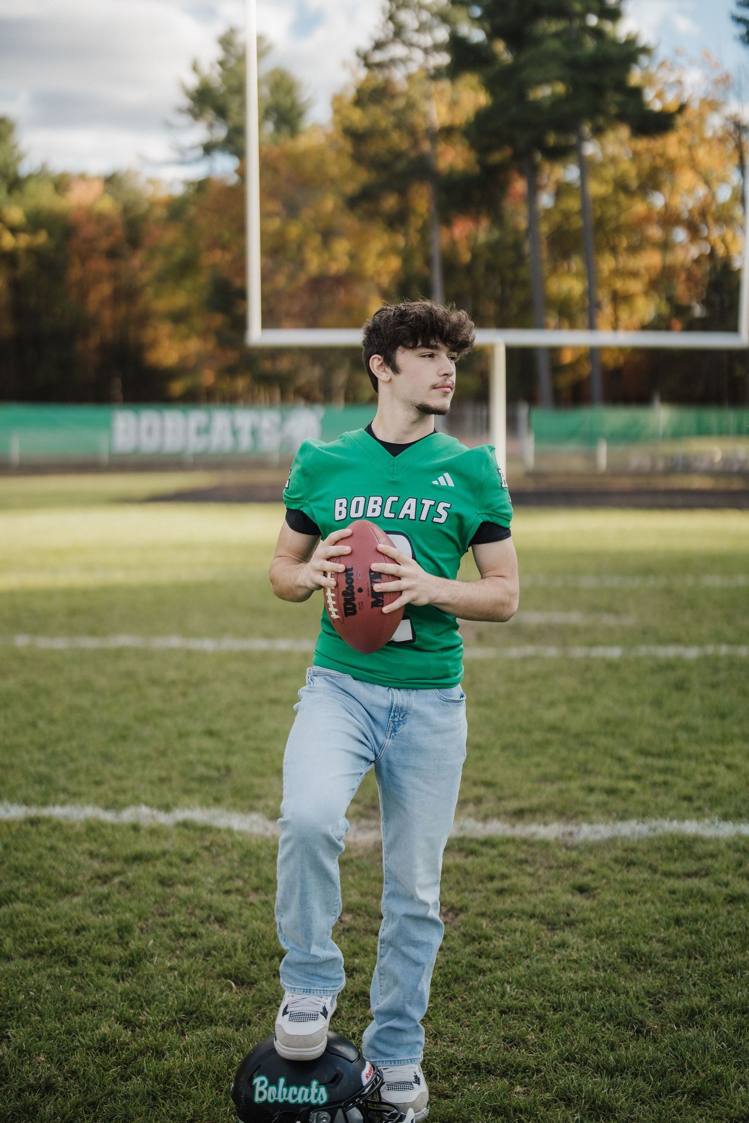 Football player in Bobcats jersey and jeans poses with ball on autumn field during practice session.