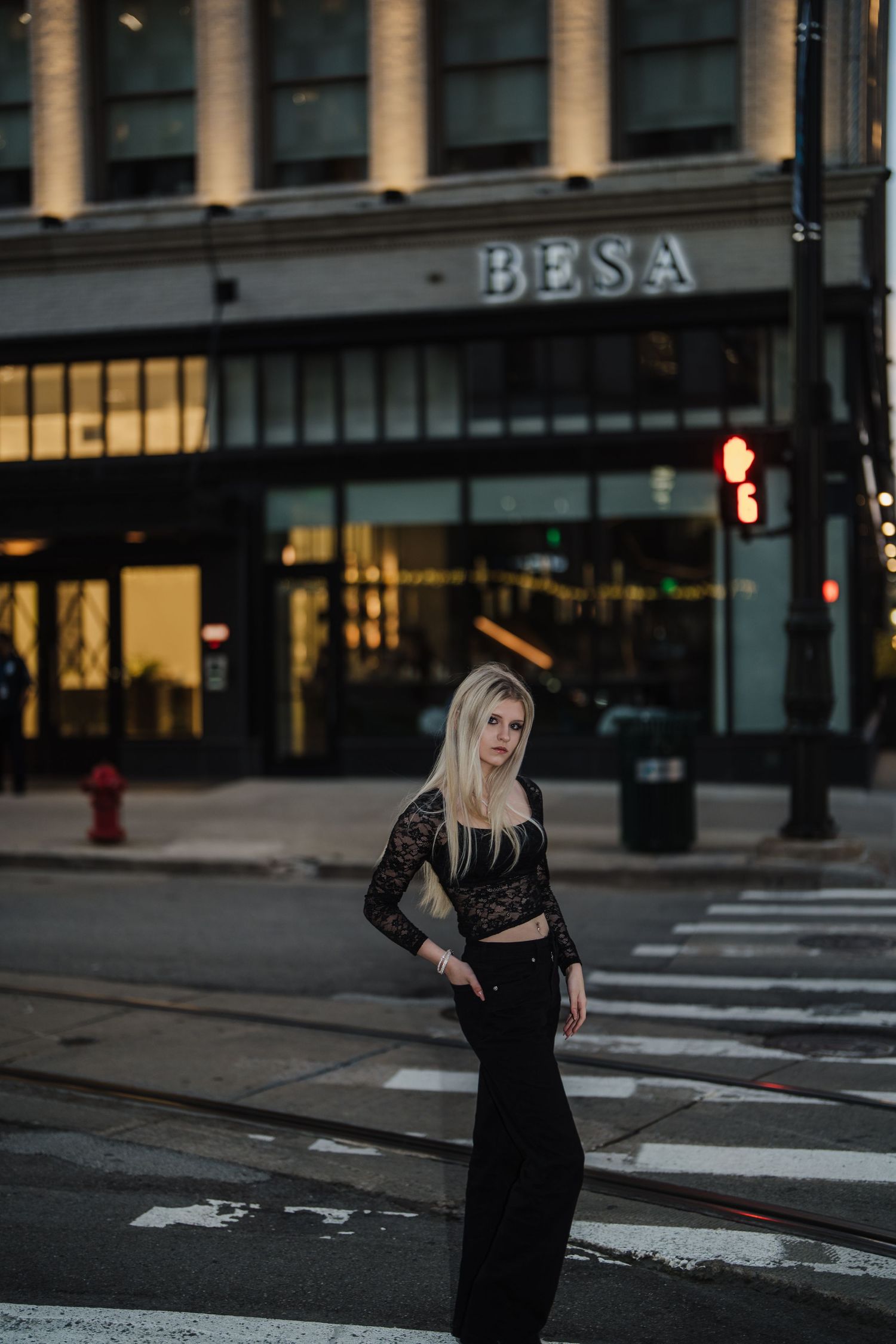 A person in a black dress stands on a city street at dusk with illuminated storefronts in the background.