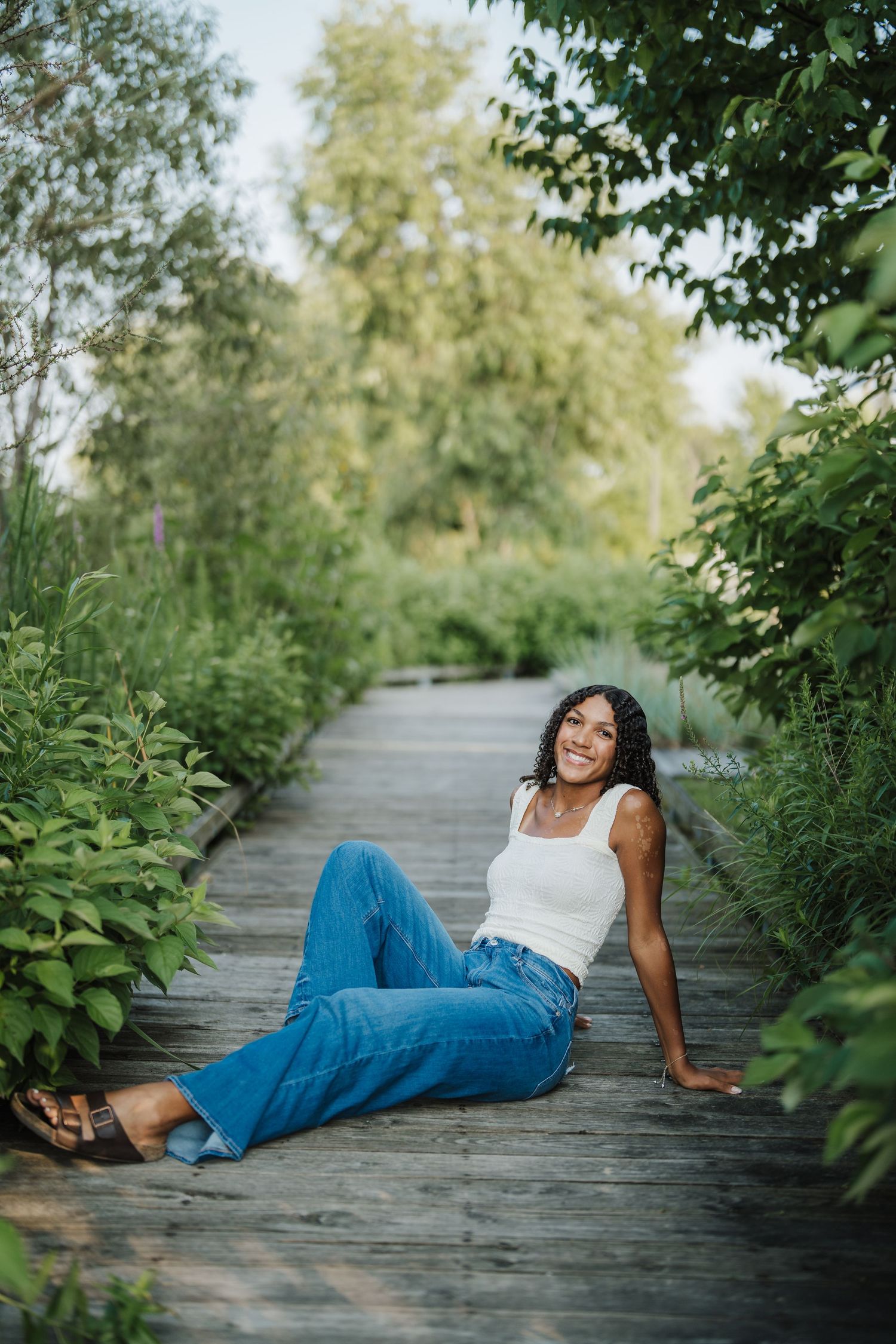 A person in a white top and blue jeans poses casually on a wooden boardwalk surrounded by lush greenery.