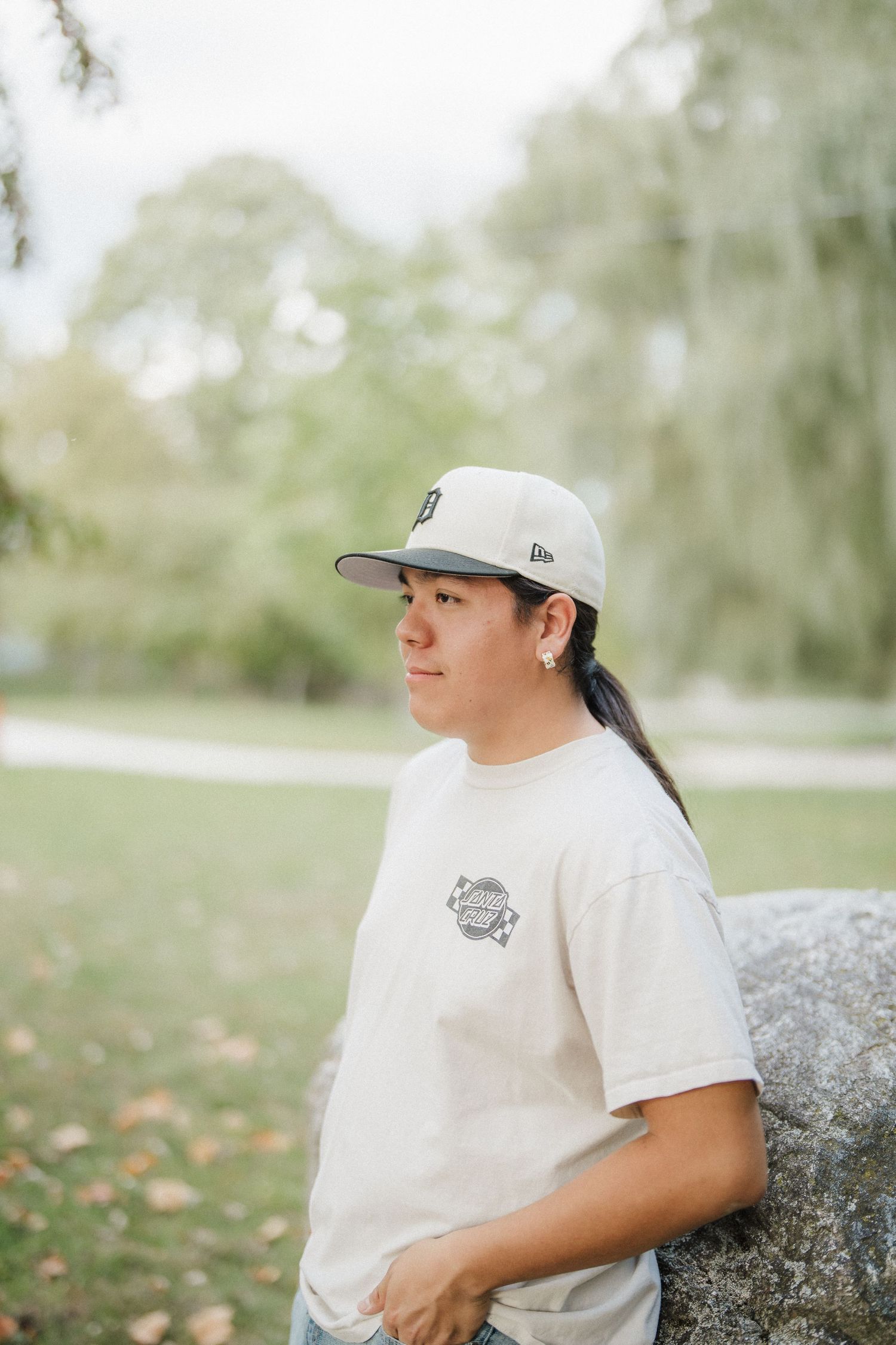A person in a white t-shirt and baseball cap sits outdoors on a sunny day with trees in the background.