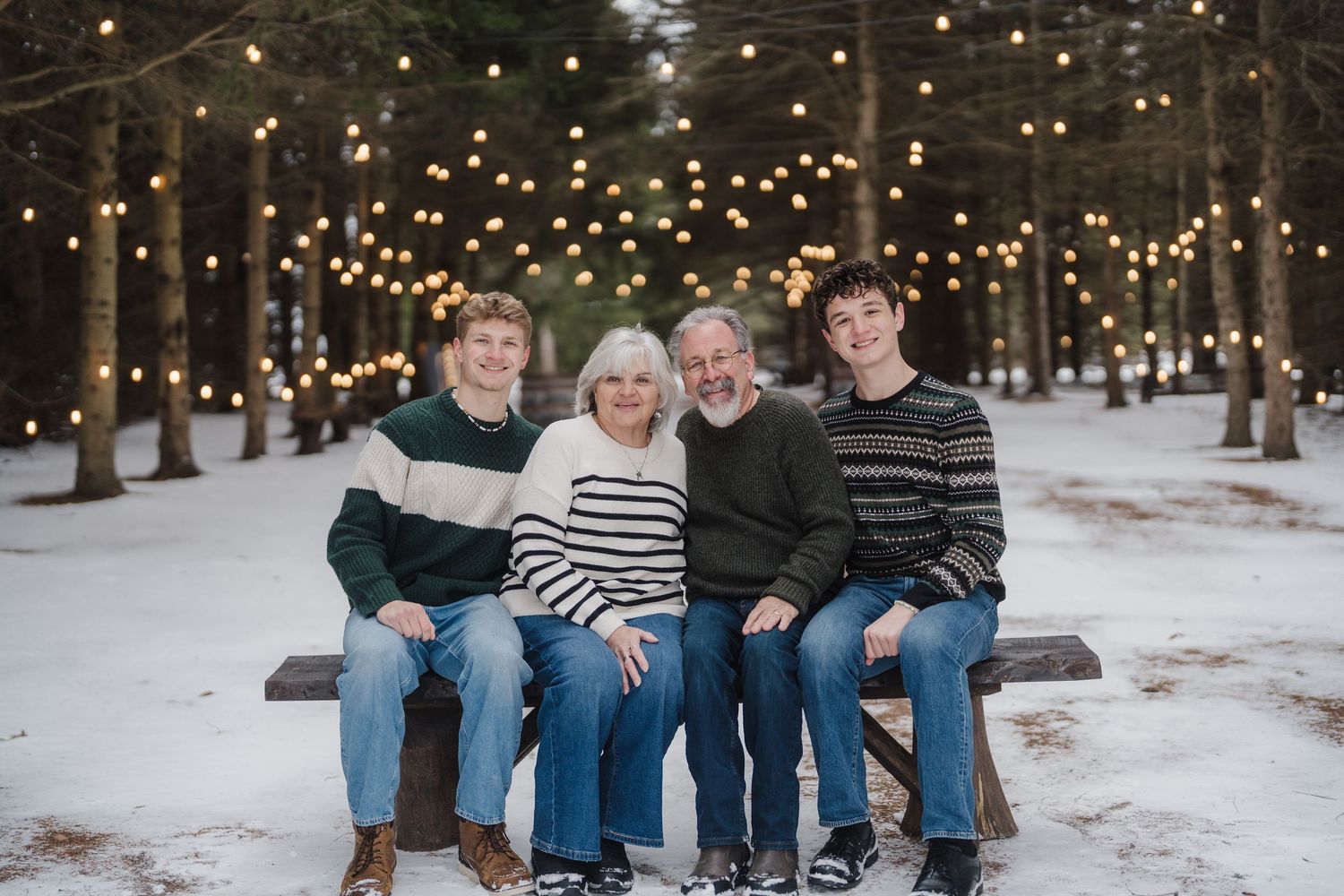 A group sits together on a wooden bench in a snowy forest setting with twinkling string lights overhead.