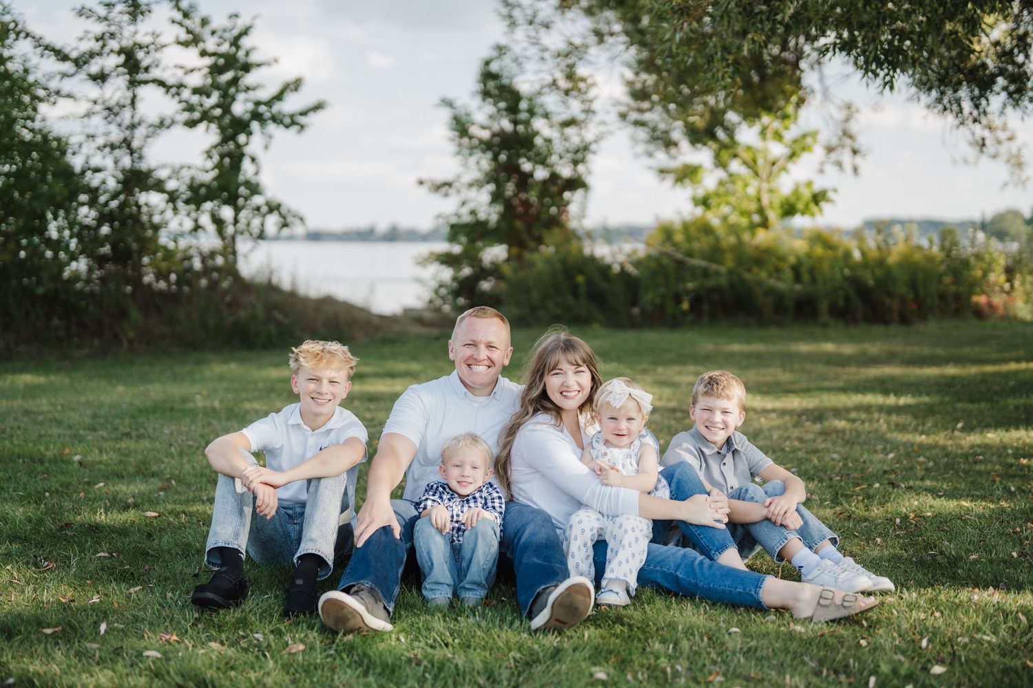 Family wearing coordinated blue and white outfits relaxes together on grassy hill during outdoor photo session.