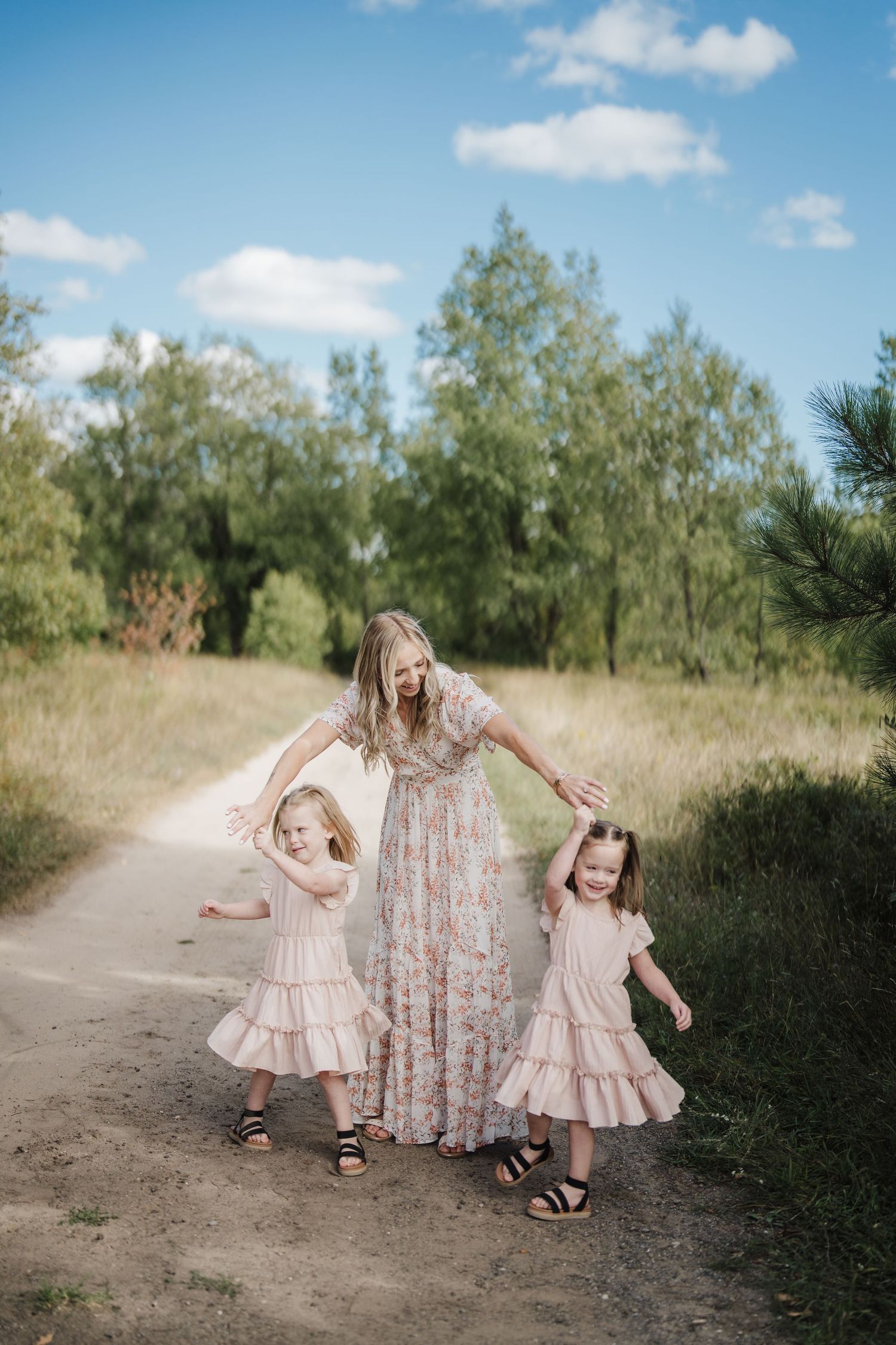 A mother and two young children in floral dresses dance together on a dirt path surrounded by green foliage.