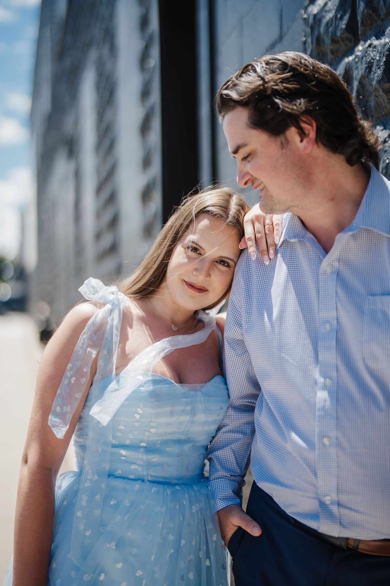 A romantic couple in light summer attire share an intimate moment outdoors against an urban backdrop on a sunny day.