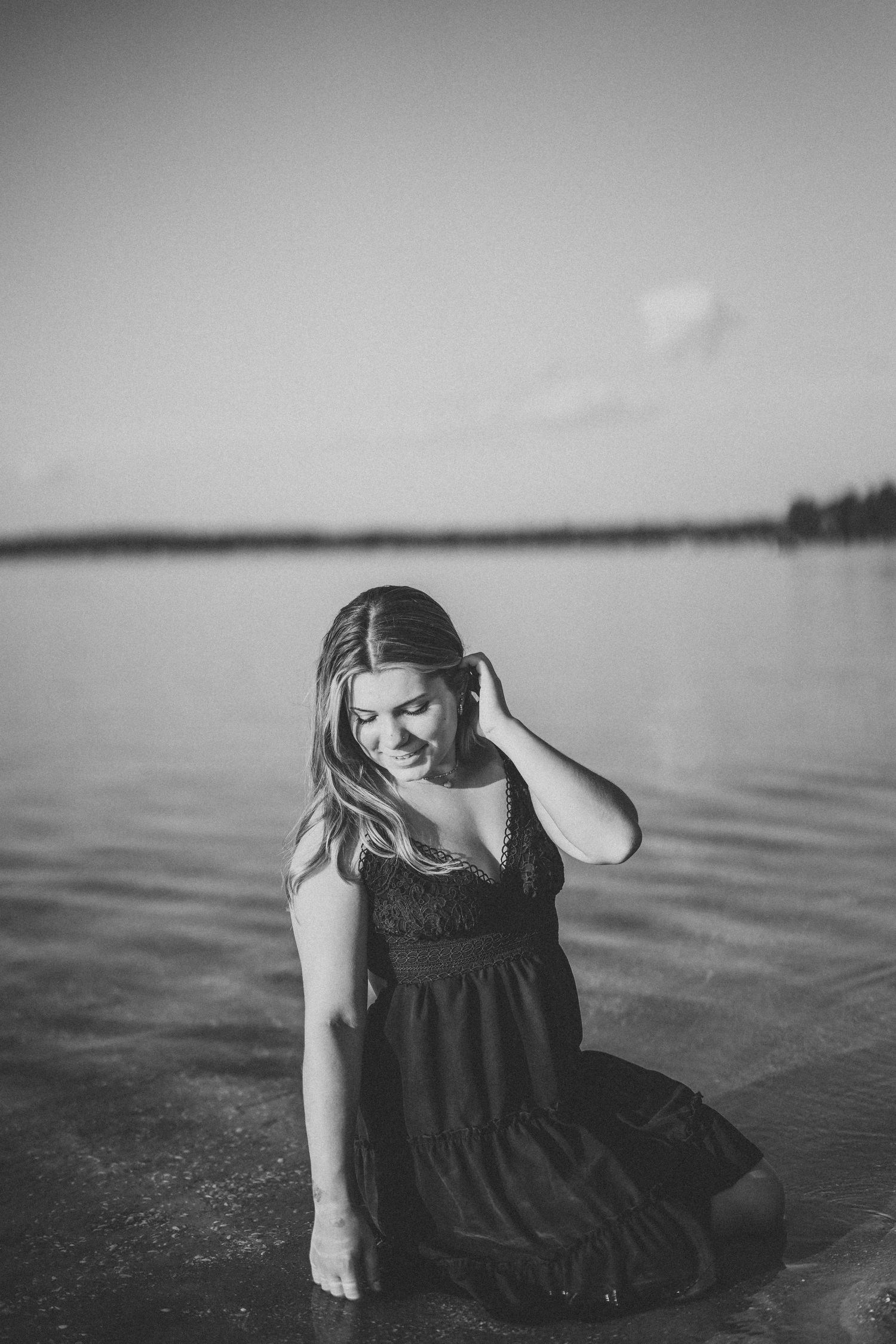 A person in a dark dress sits by a serene lake in a moody black and white photograph.