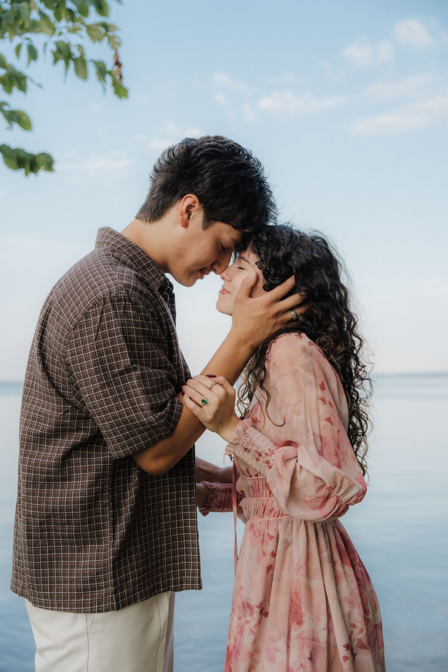 A romantic couple sharing an intimate moment by the water, holding hands and touching foreheads against a serene blue sky backdrop.