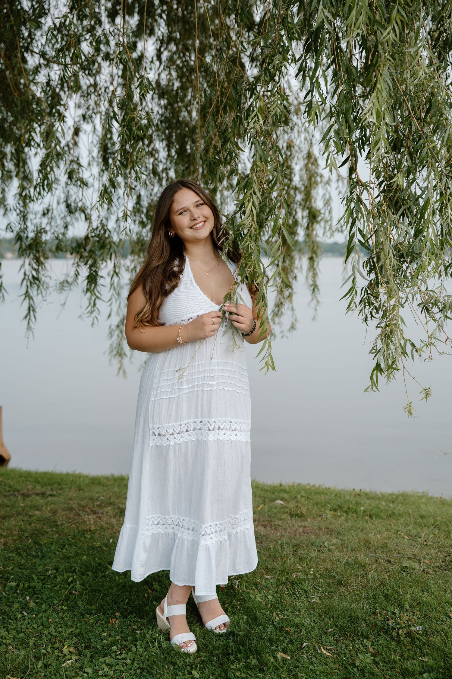 Young woman in white maxi dress smiling under weeping willow tree by a lake on a sunny day.