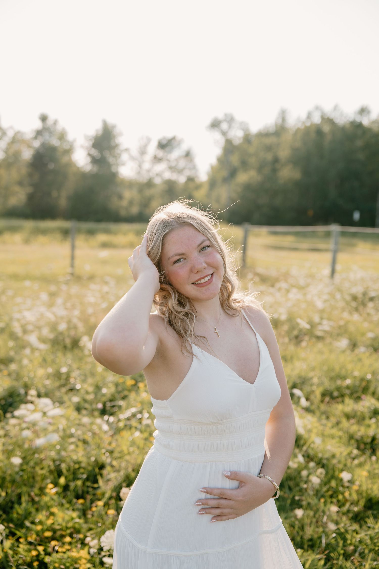 Young blonde woman in white dress smiling in sunny wildflower field with trees in background.