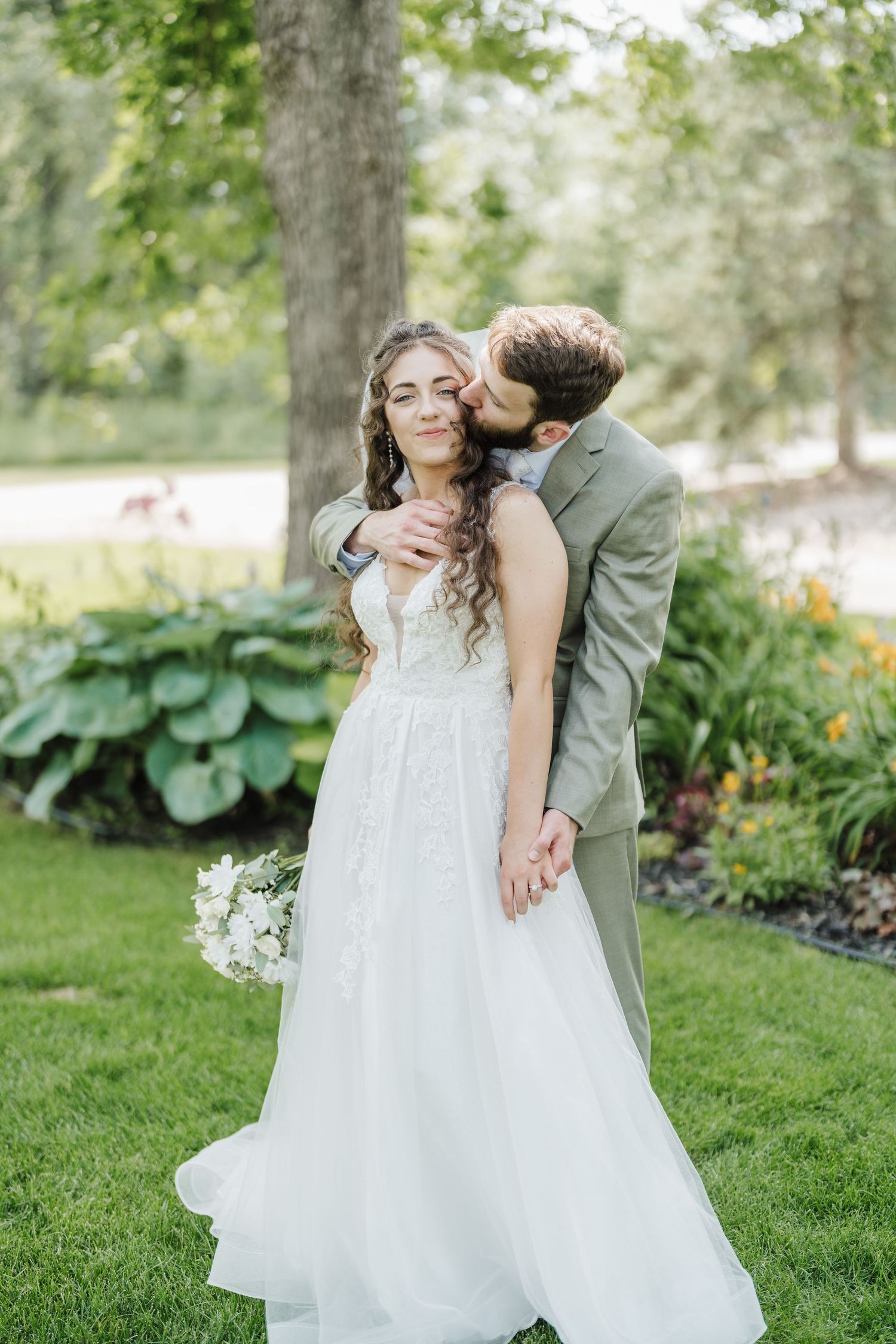 A romantic wedding couple embraces in a garden setting with the bride holding a bouquet.