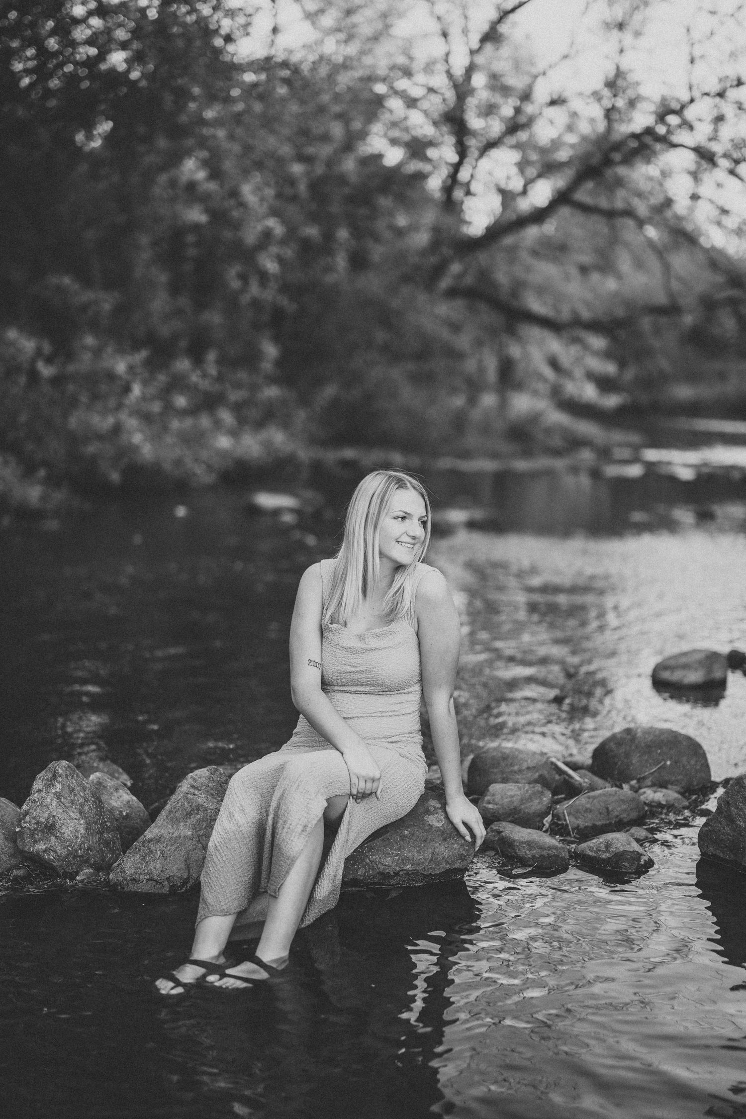 A person in a light dress sits on rocks by a tranquil river in a black and white nature scene.