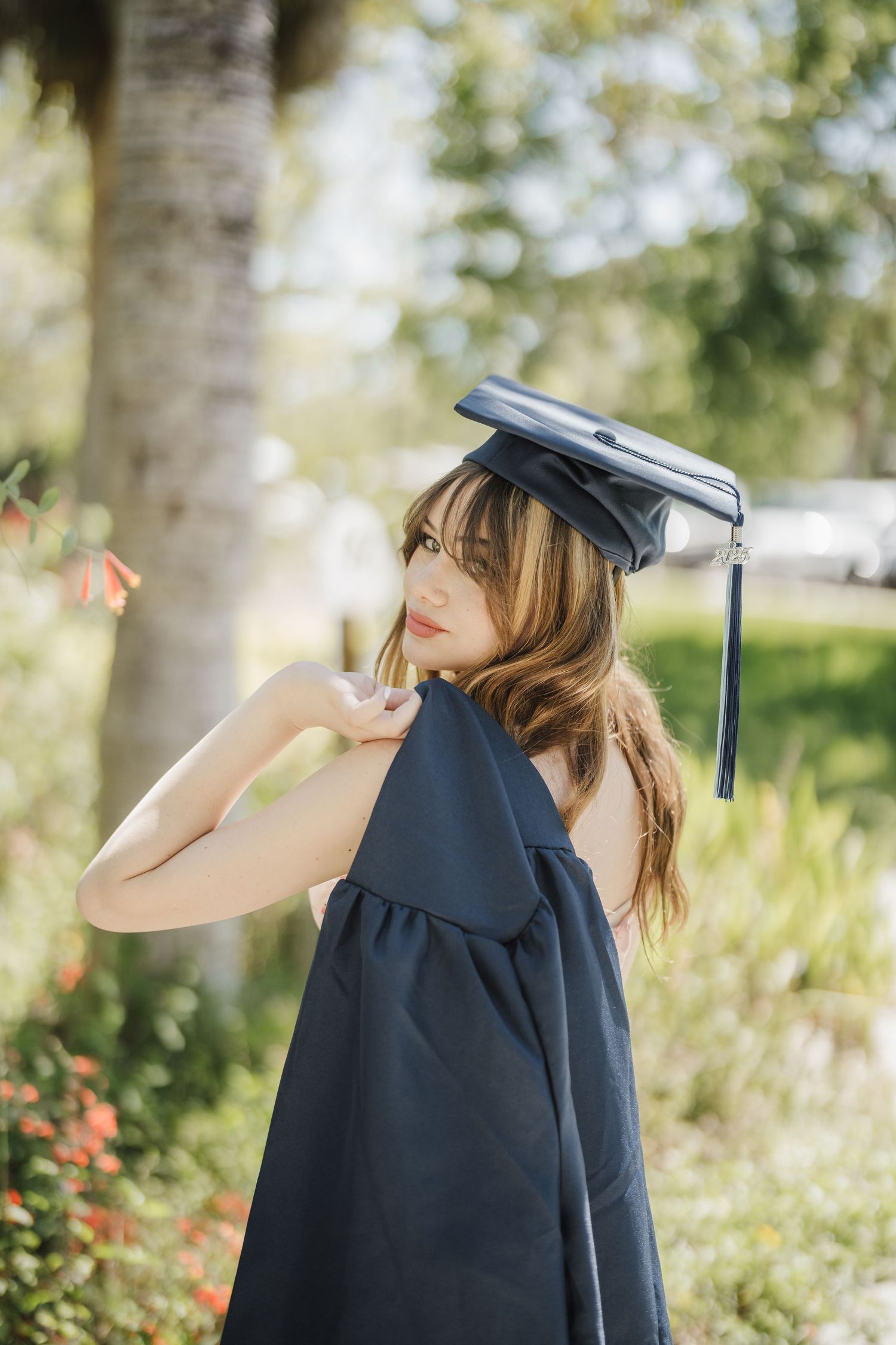 Graduate wearing black cap and gown poses thoughtfully in a sunny garden with palm trees and flowers.
