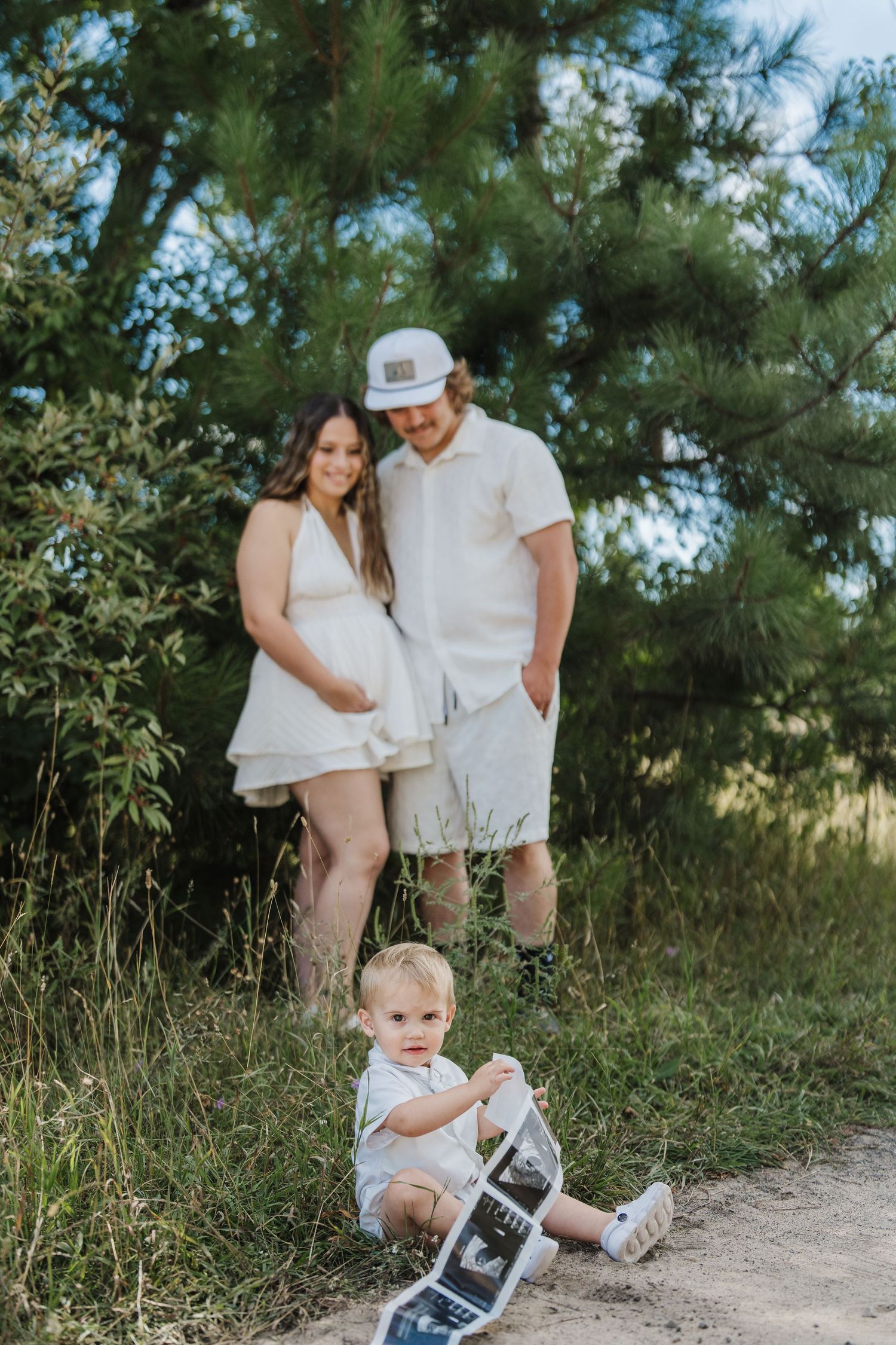 A couple in white outfits stands with their toddler on a dirt path surrounded by lush greenery and pine trees.