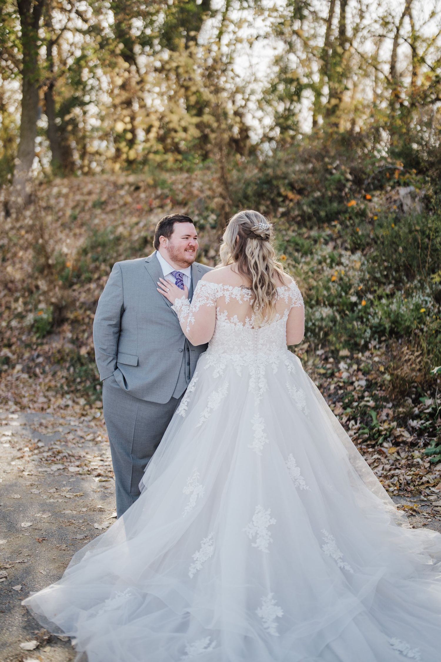Bride in flowing lace dress and groom in gray suit share a candid moment on an autumn path.