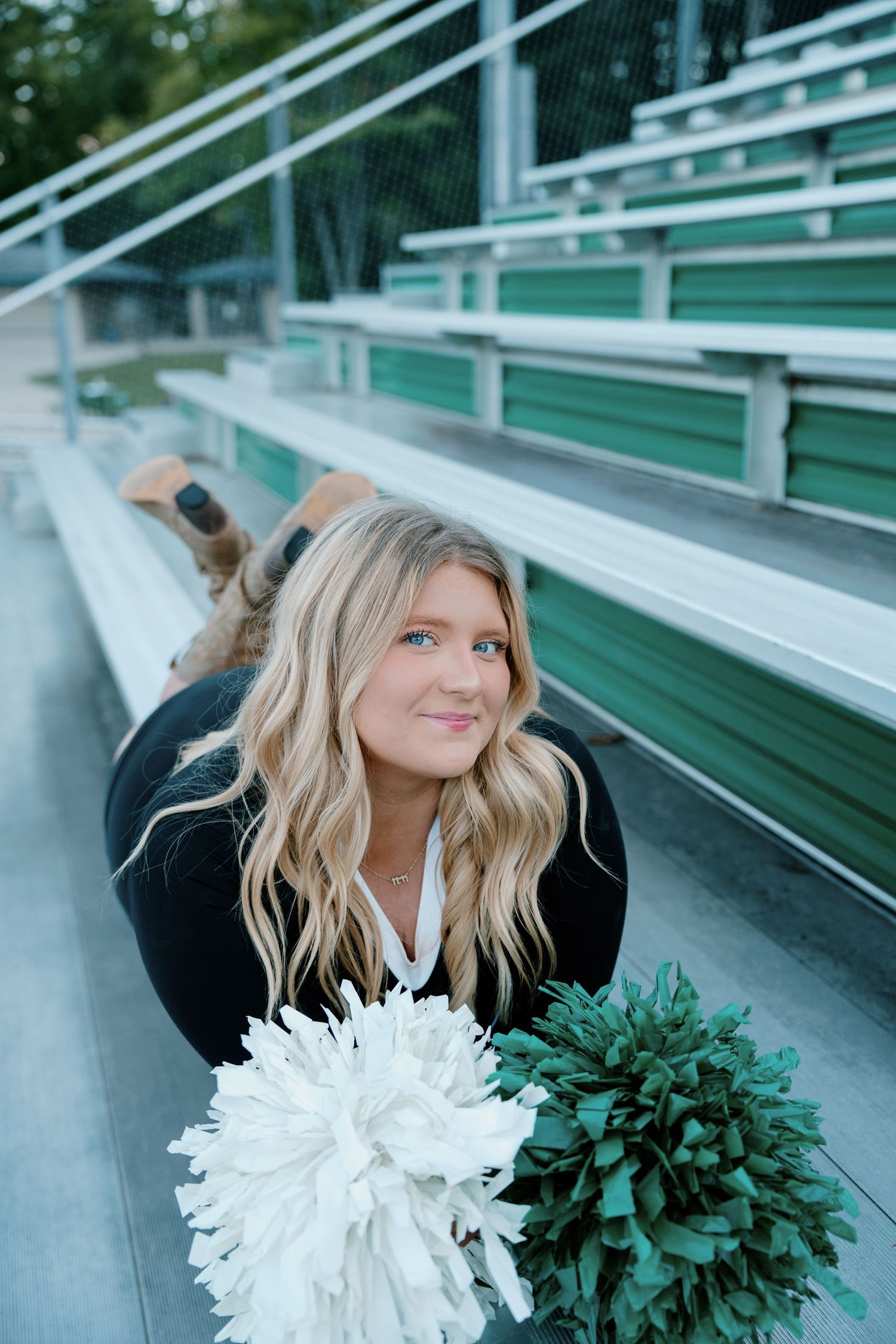 Cheerleader with white and green pom-poms posing on bleachers with long blonde wavy hair.