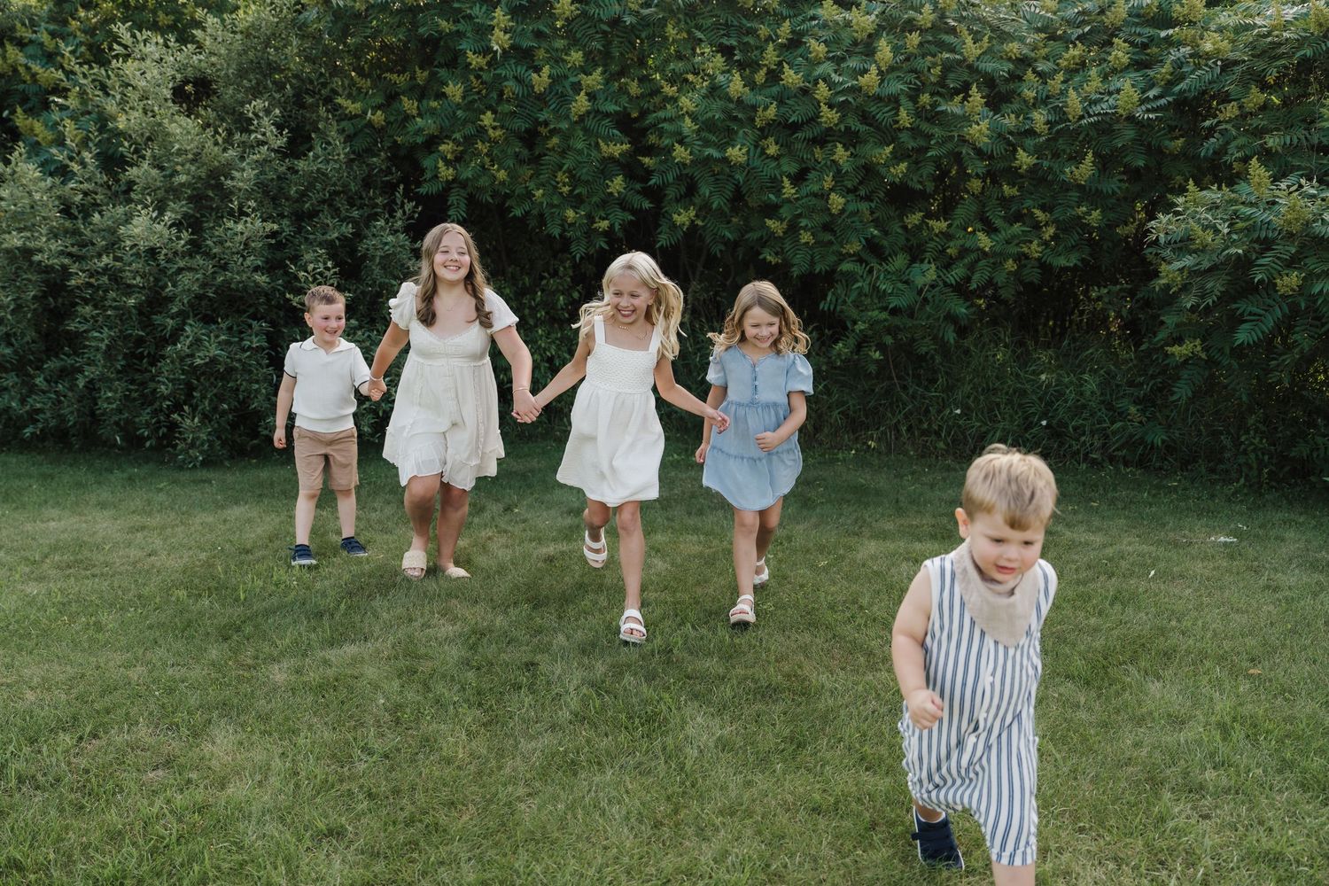 Five children in summer outfits run and hold hands across a green lawn with lush bushes in the background.