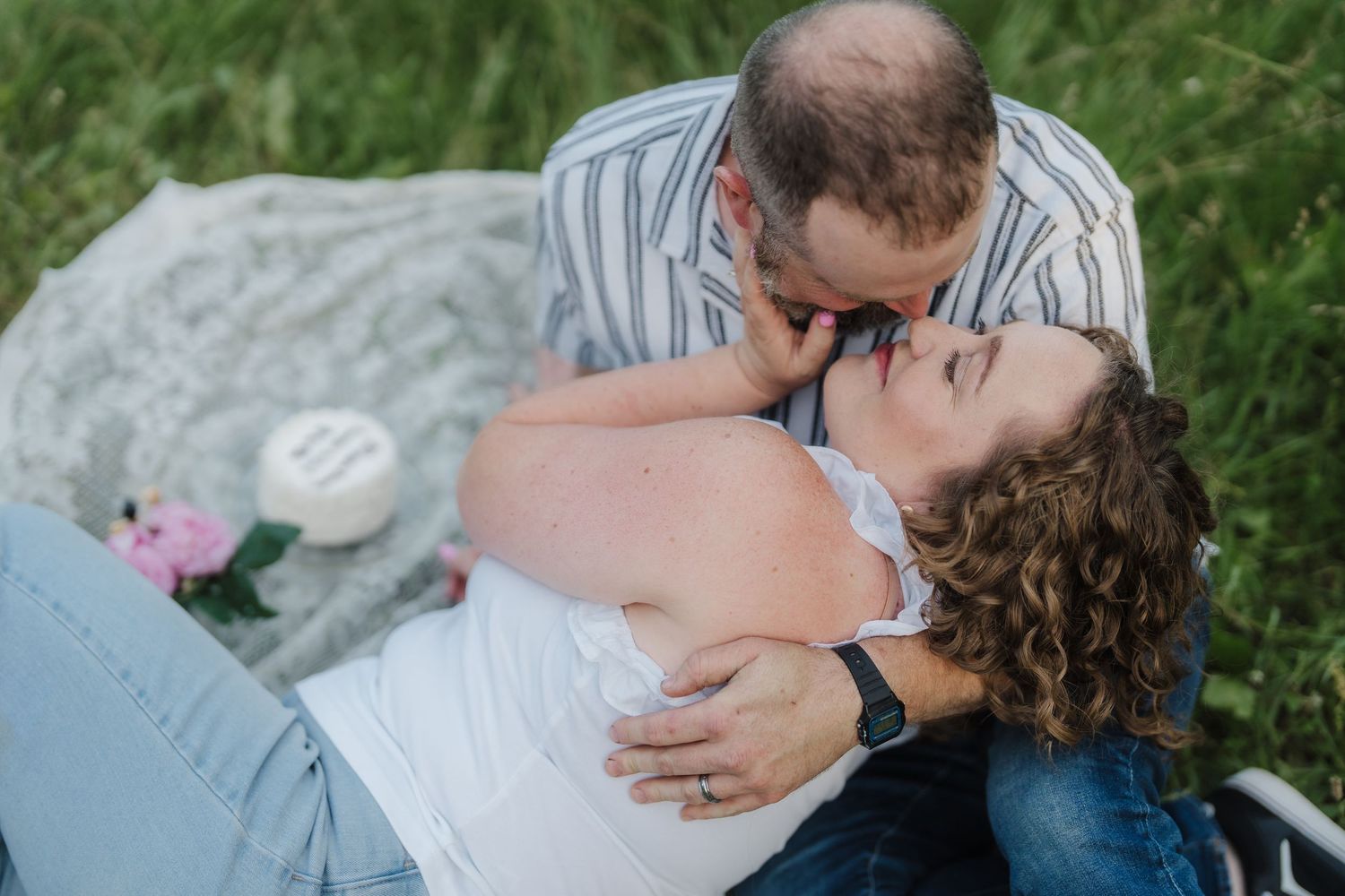 Couple sharing a romantic kiss while lying on a blanket in a lush green field outdoors.