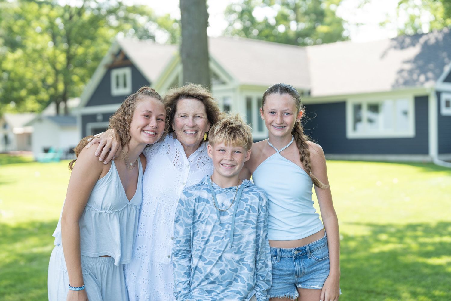 A smiling family of four poses outdoors on green grass in front of a dark blue house on a sunny day.