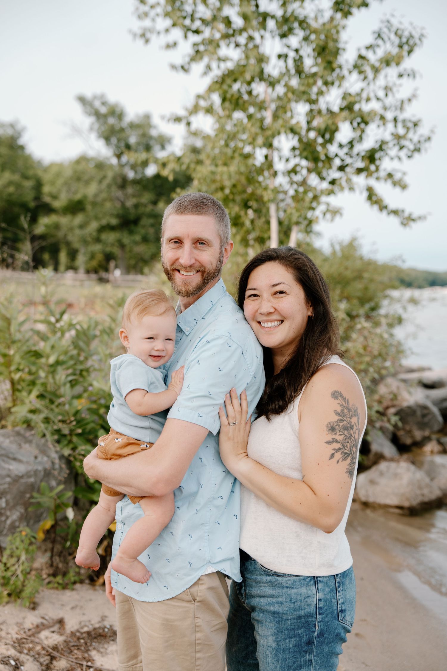 A smiling family of three poses outdoors near a rocky shoreline, with a man holding a baby and a woman beside them.