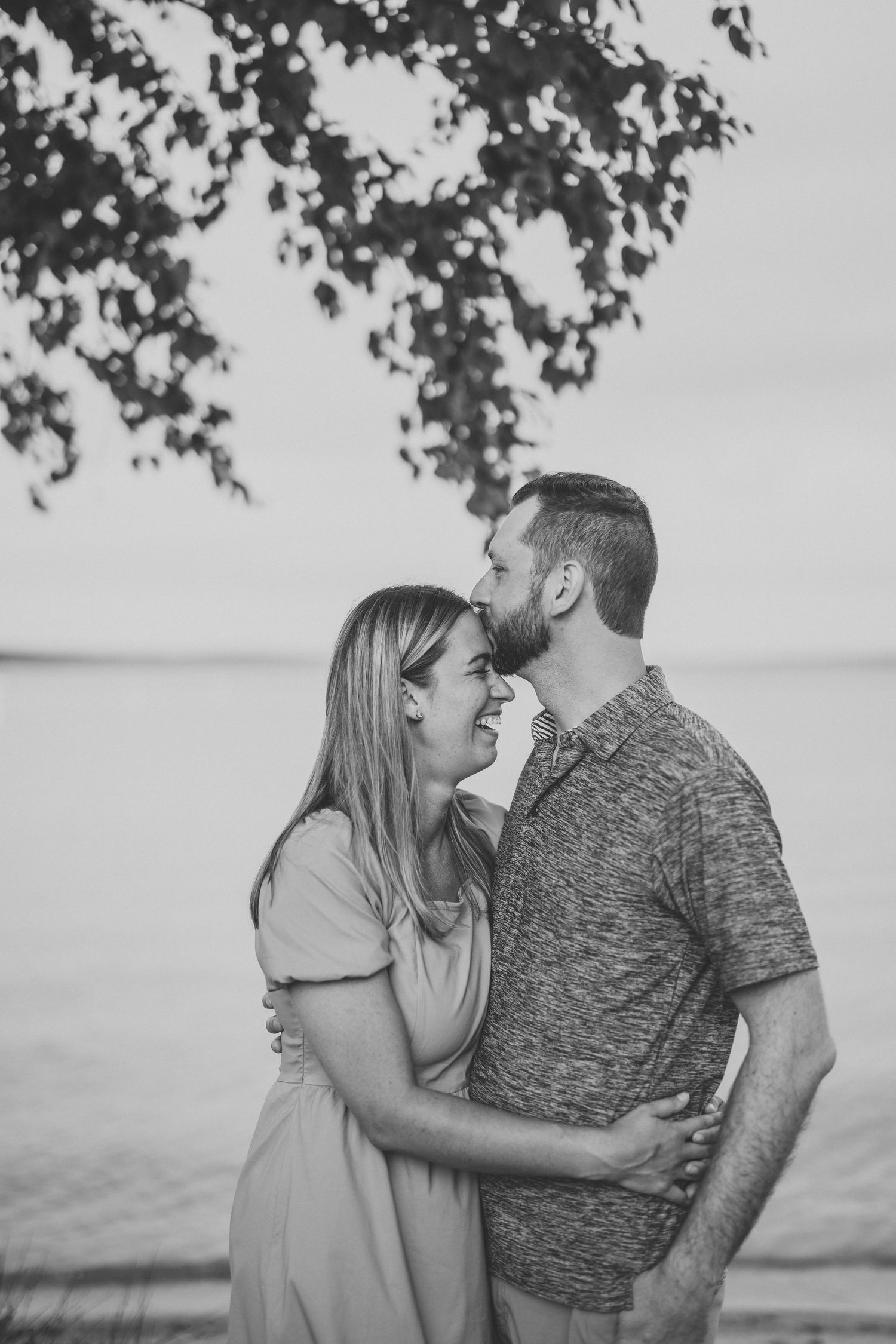 Black and white photo of couple embracing and laughing together outdoors under tree branches.