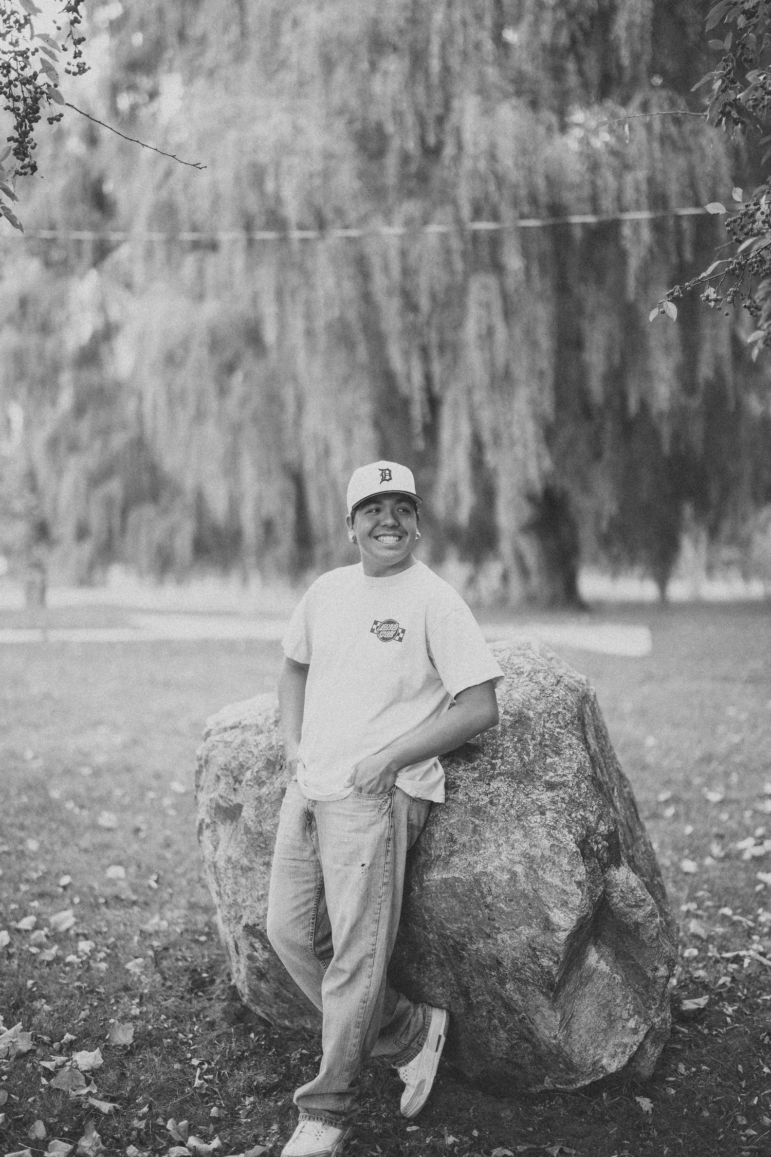 Black and white photo of someone in casual wear leaning against a large boulder in a park setting with willow trees.