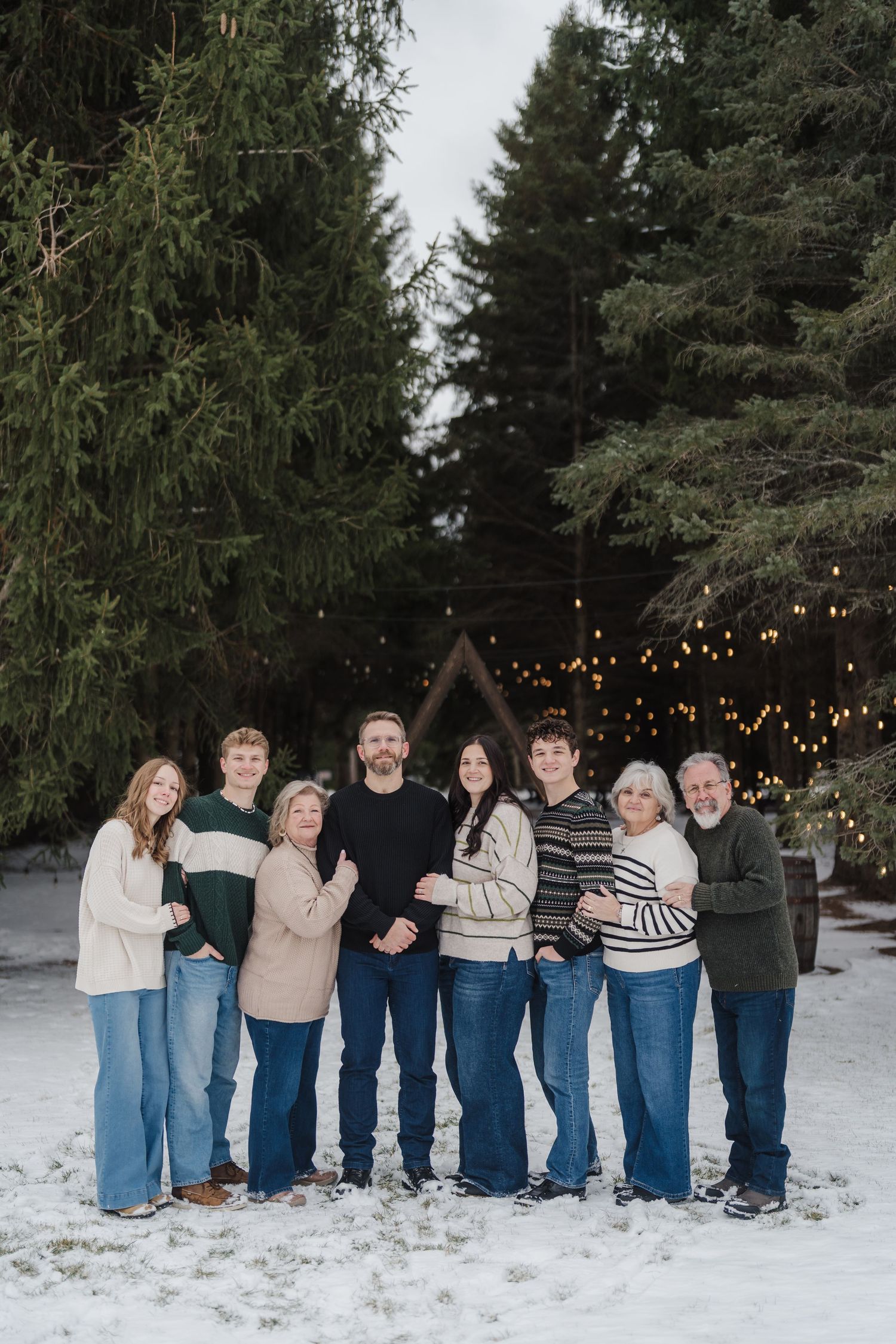 A group wearing coordinated blue jeans and light tops stands together in a snowy winter setting with string lights behind them.