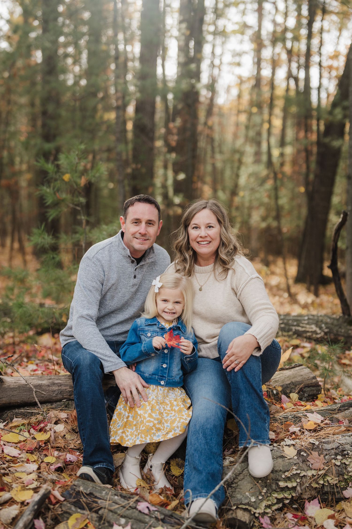 A family sits together on fallen logs in an autumn forest setting during a casual outdoor photo session.
