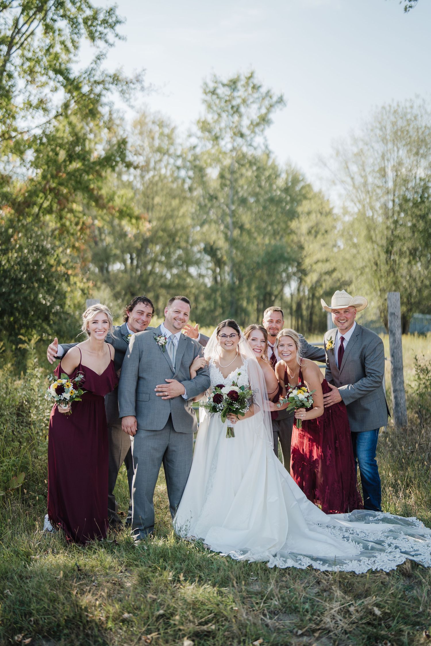 Wedding party in burgundy and gray attire poses together outdoors in a natural setting during autumn.