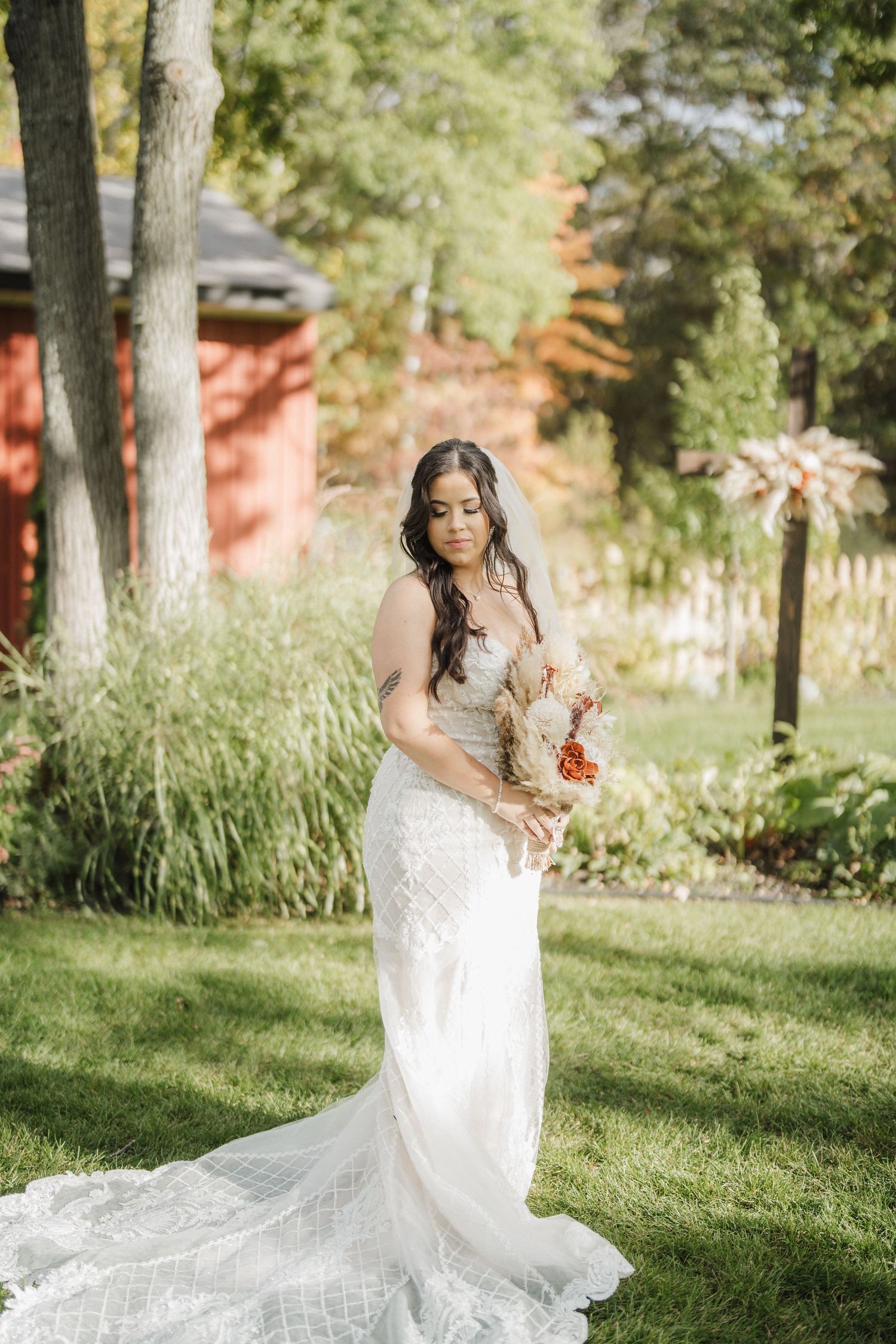 Bride in flowing lace gown poses gracefully on sunlit grass with trees in background.