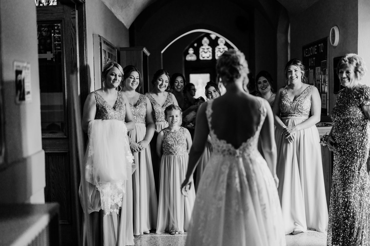 A black and white photo of bridesmaids in long dresses gathered in a hallway awaiting the bride.