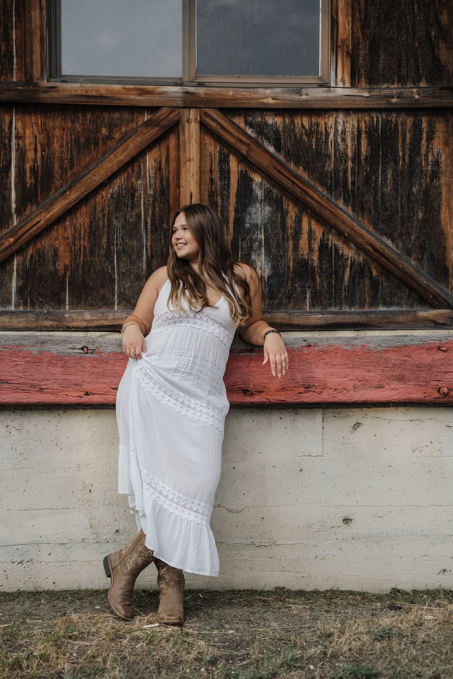 Young woman in white maxi dress and brown cowboy boots leaning against rustic barn wall, smiling outdoors.
