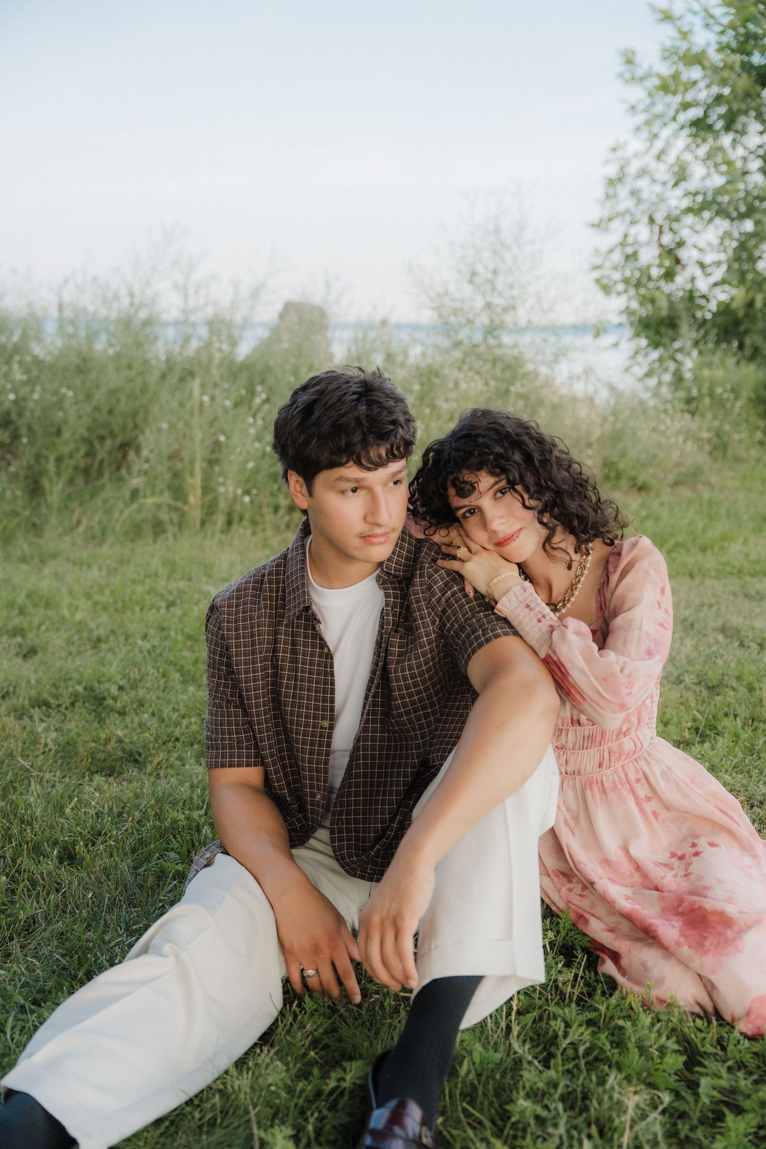 A young couple sitting together on green grass outdoors, woman in pink dress leaning on man in brown jacket.