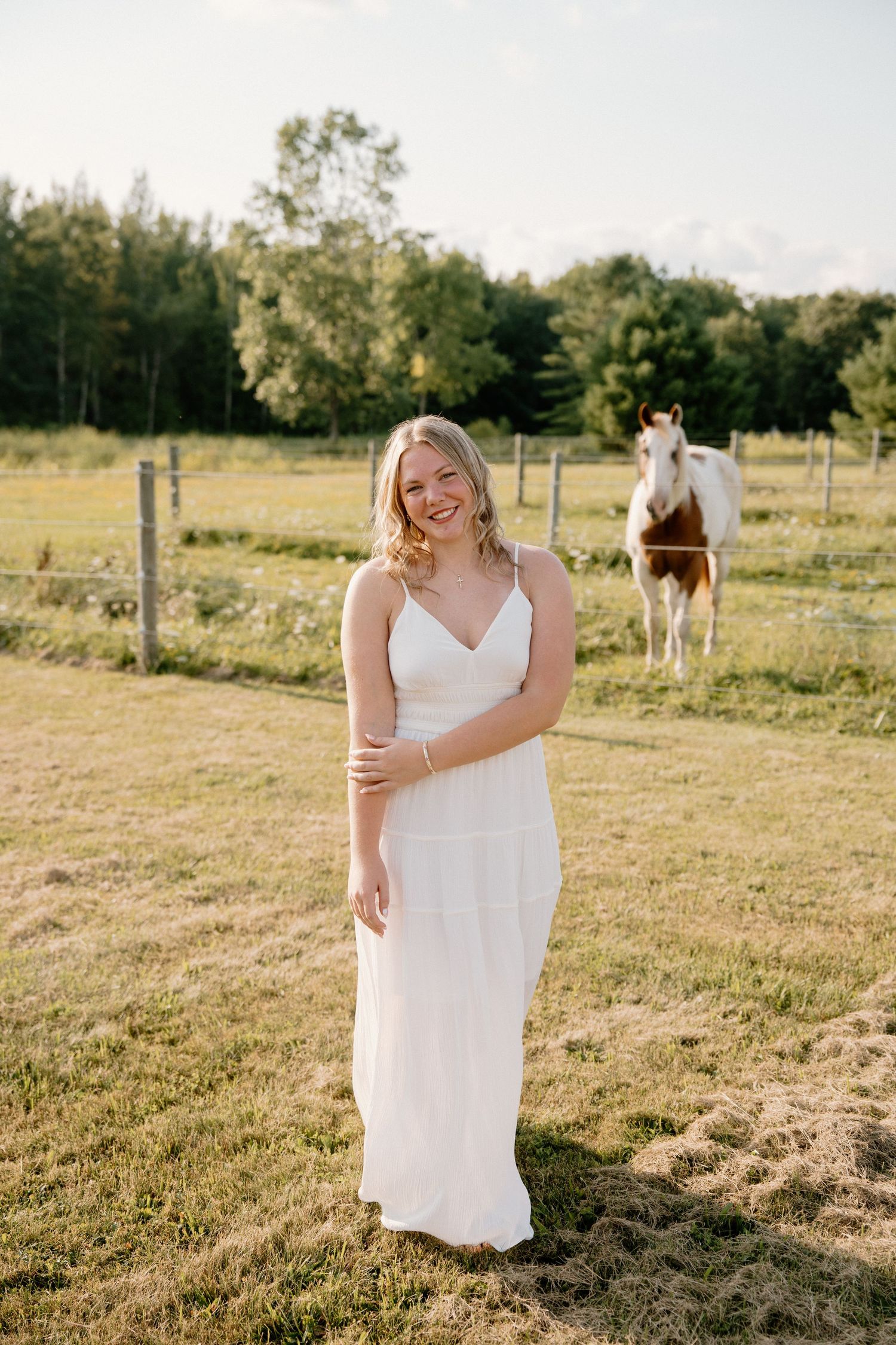 Young woman in white maxi dress smiling in sunny farm field with horse and green trees in background.