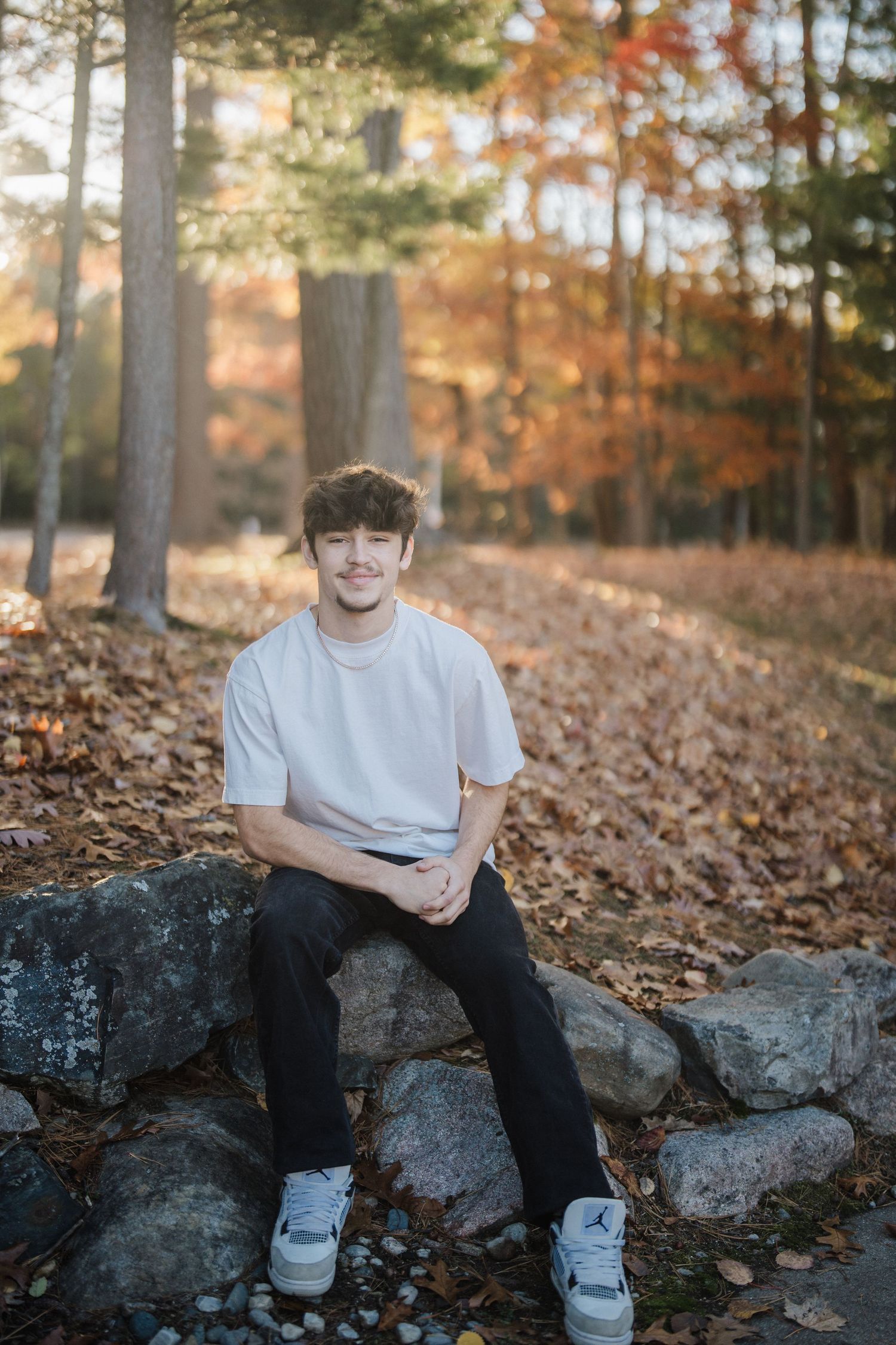 Person sitting on large rocks surrounded by fallen autumn leaves in a wooded setting.