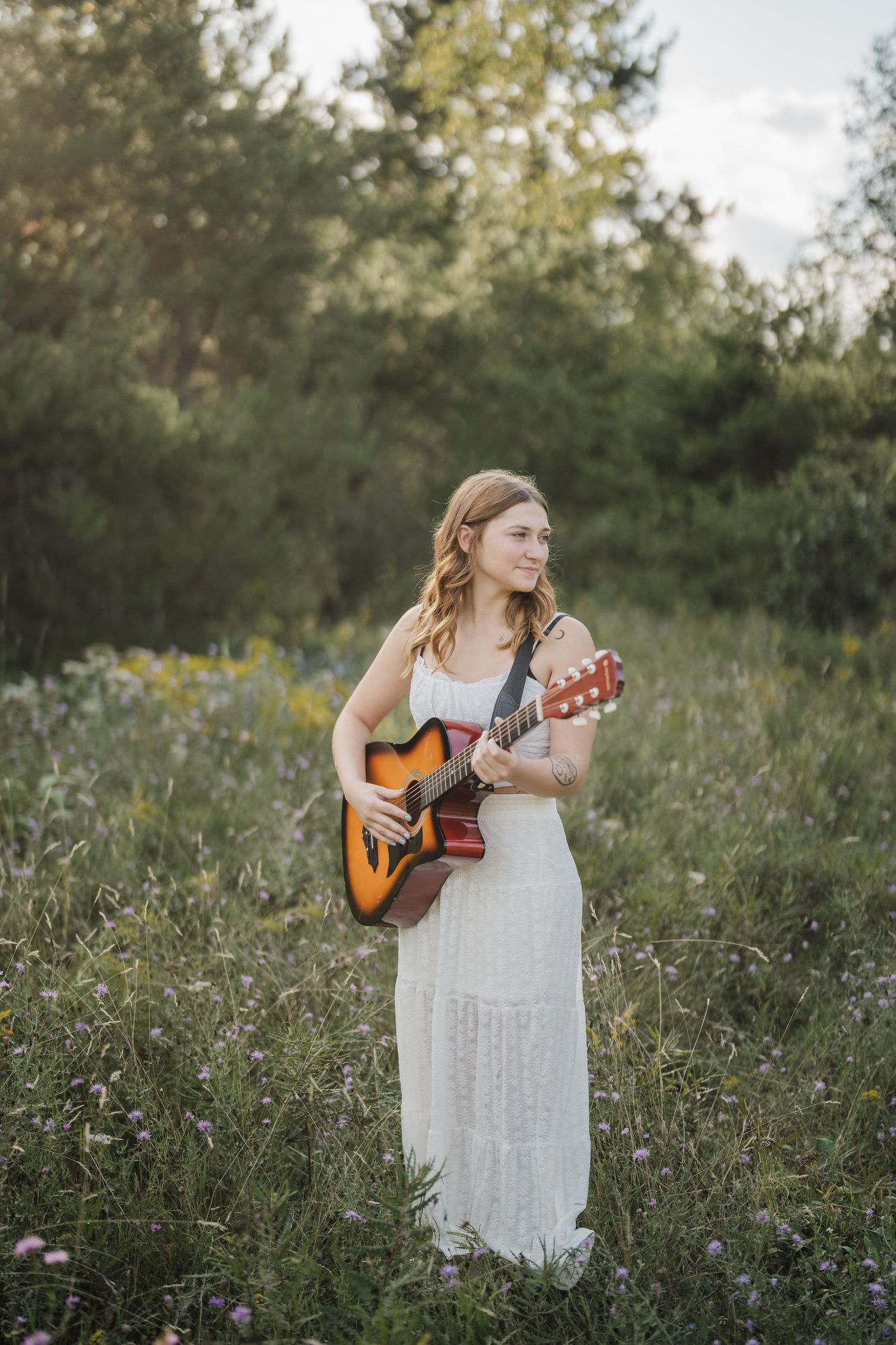 A woman in a white dress plays acoustic guitar while standing in a wildflower meadow at sunset.