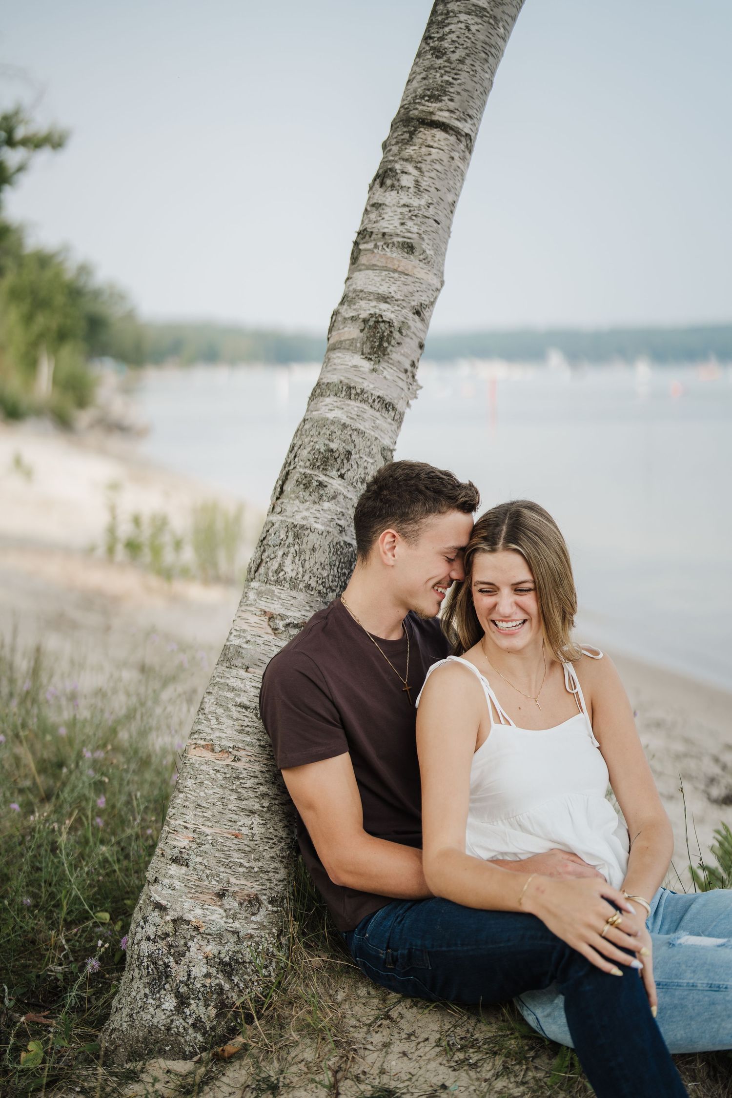 Romantic couple embraces while sitting against a birch tree trunk on a peaceful beach at sunset.